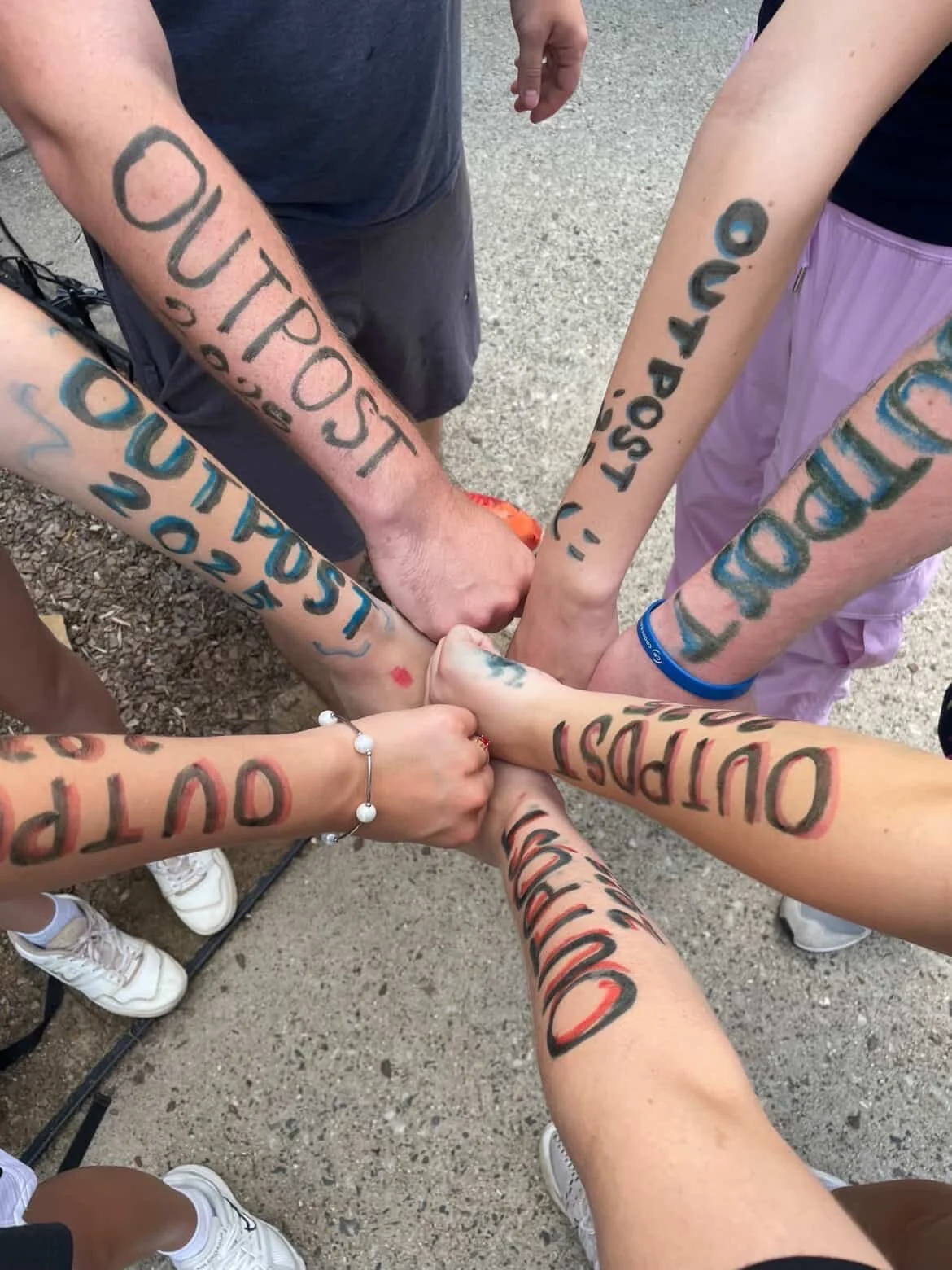 Group of people with arms extended and hands stacked together in the center, each arm with colorful tattoos and some with date and location text.