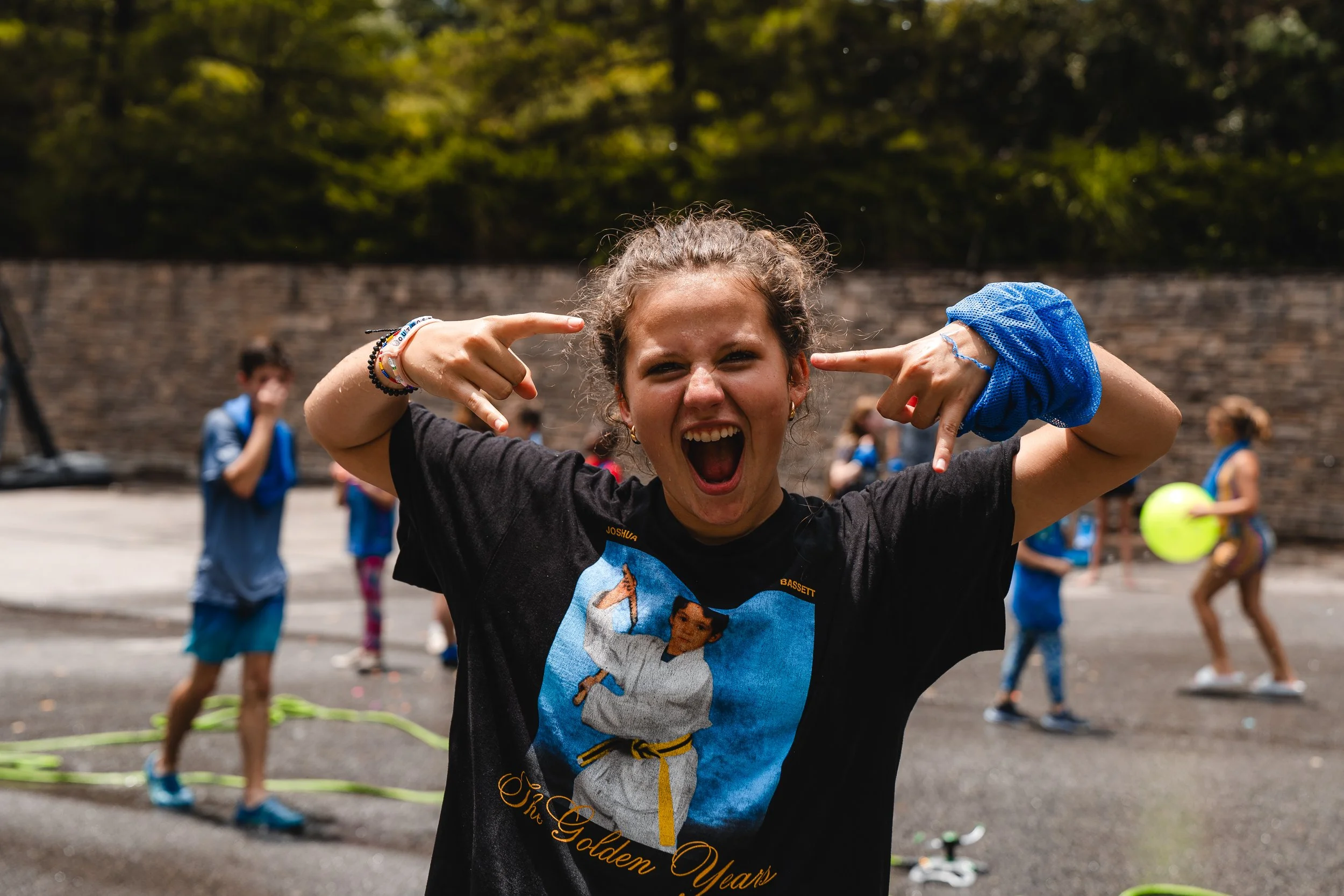 A young woman smiling and making a rock on gesture with her hands, in an outdoor setting with children playing in the background.
