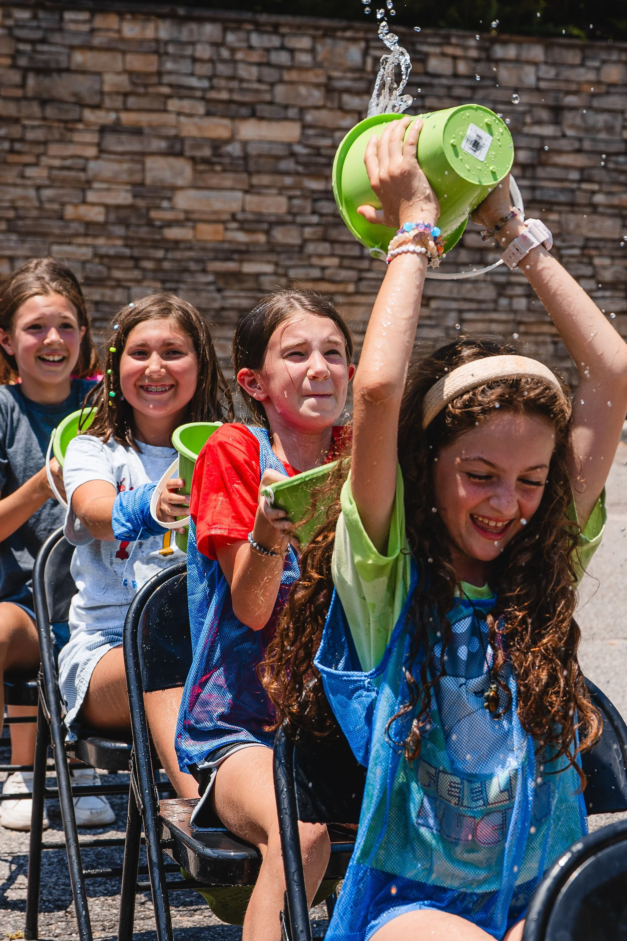 Group of girls participating in a water relay race, pouring water from buckets onto each other outside on a sunny day.
