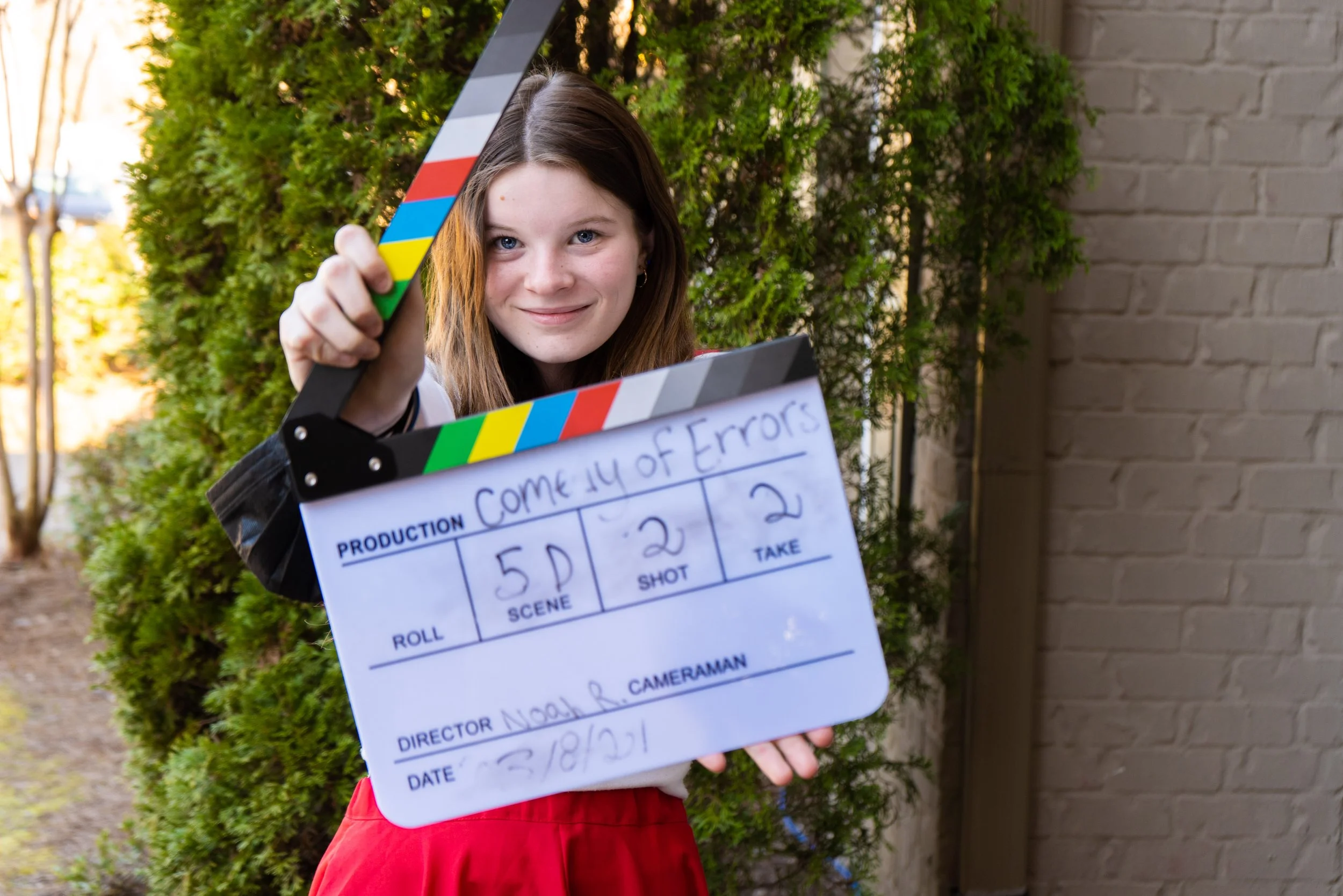 Young woman holding a film slate with 'Come Fly with Errors' written on it, standing outside near greenery and a brick wall.