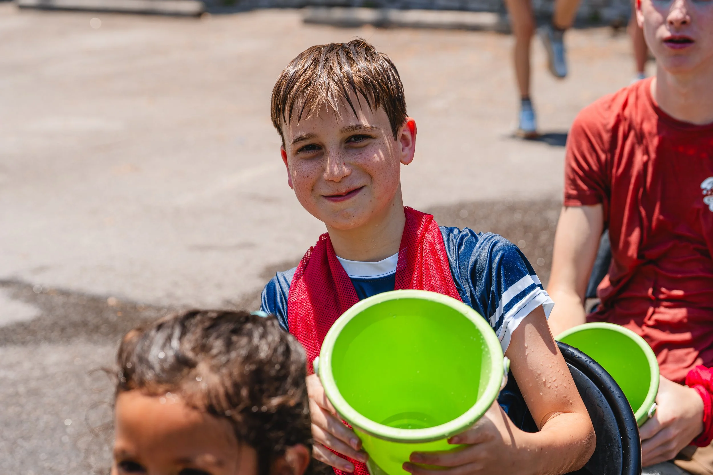 A boy with wet skin and freckles holding a bright green bucket, smiling at the camera outdoors under sunlight, with other kids in the background.