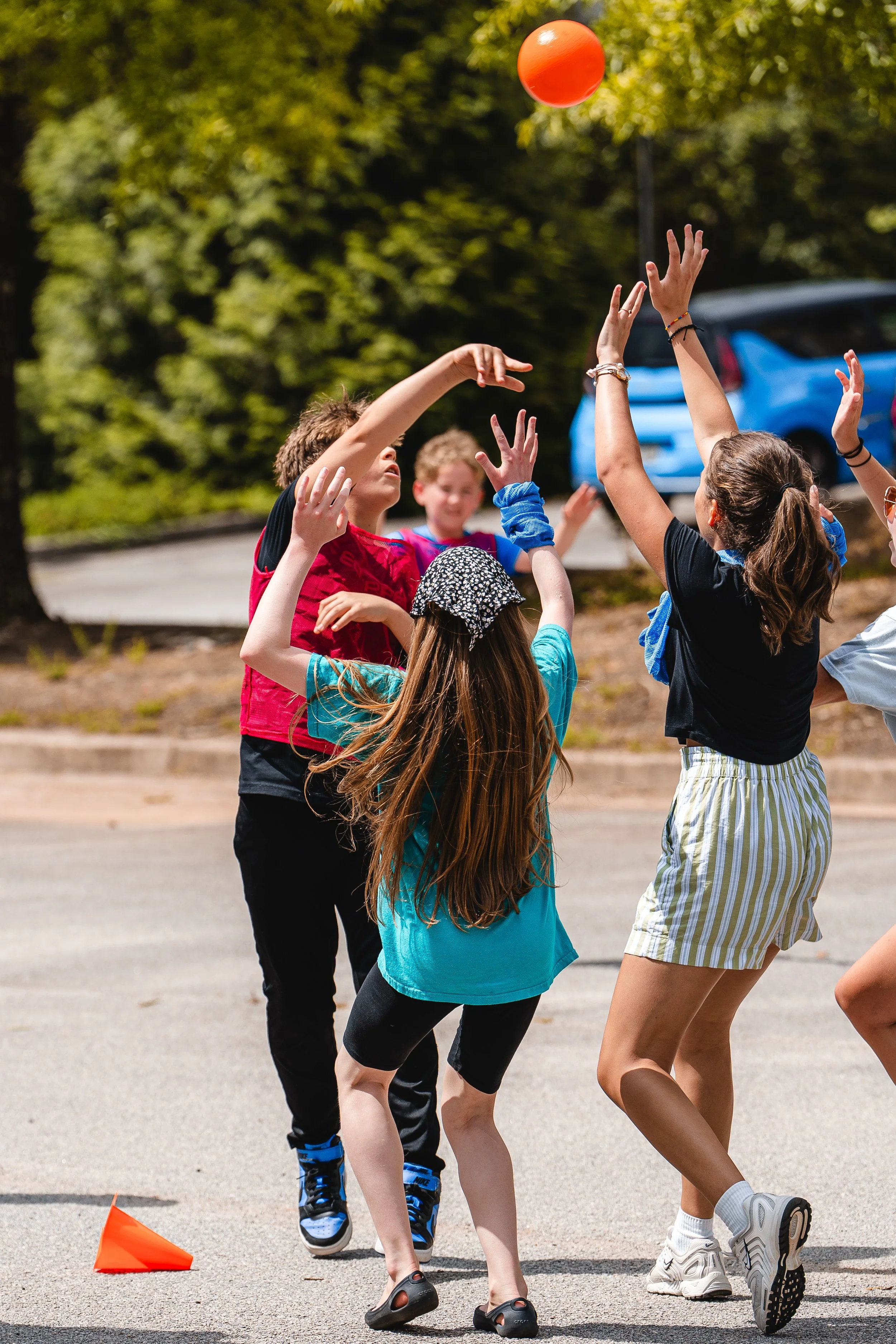 Children playing outdoor volleyball, jumping to hit an orange ball, with a traffic cone on the ground, in a parking lot with trees and a blue car in the background.