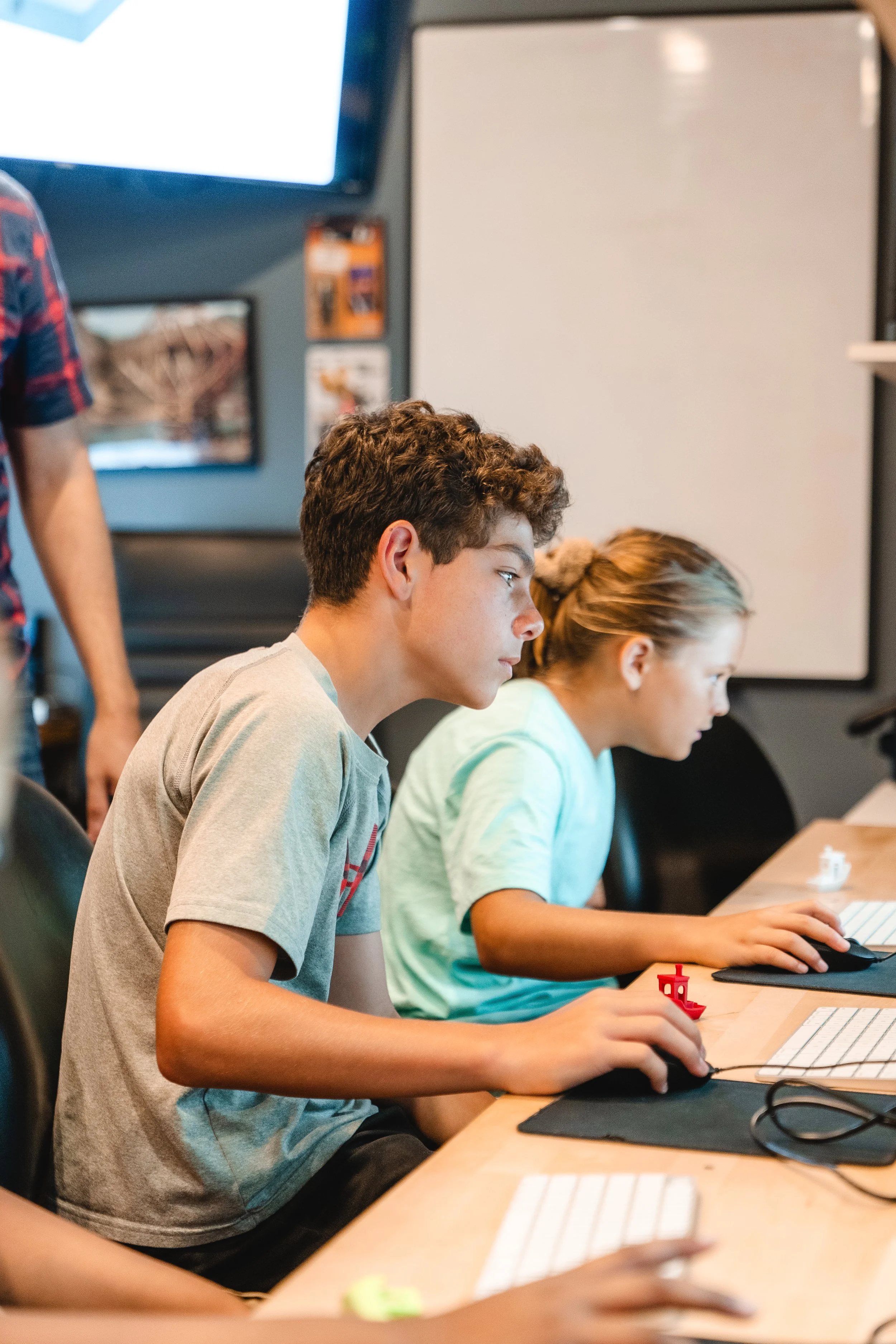 Two kids, a boy and a girl, are sitting at a desk, using computers and focusing on their screens in a classroom or computer lab.