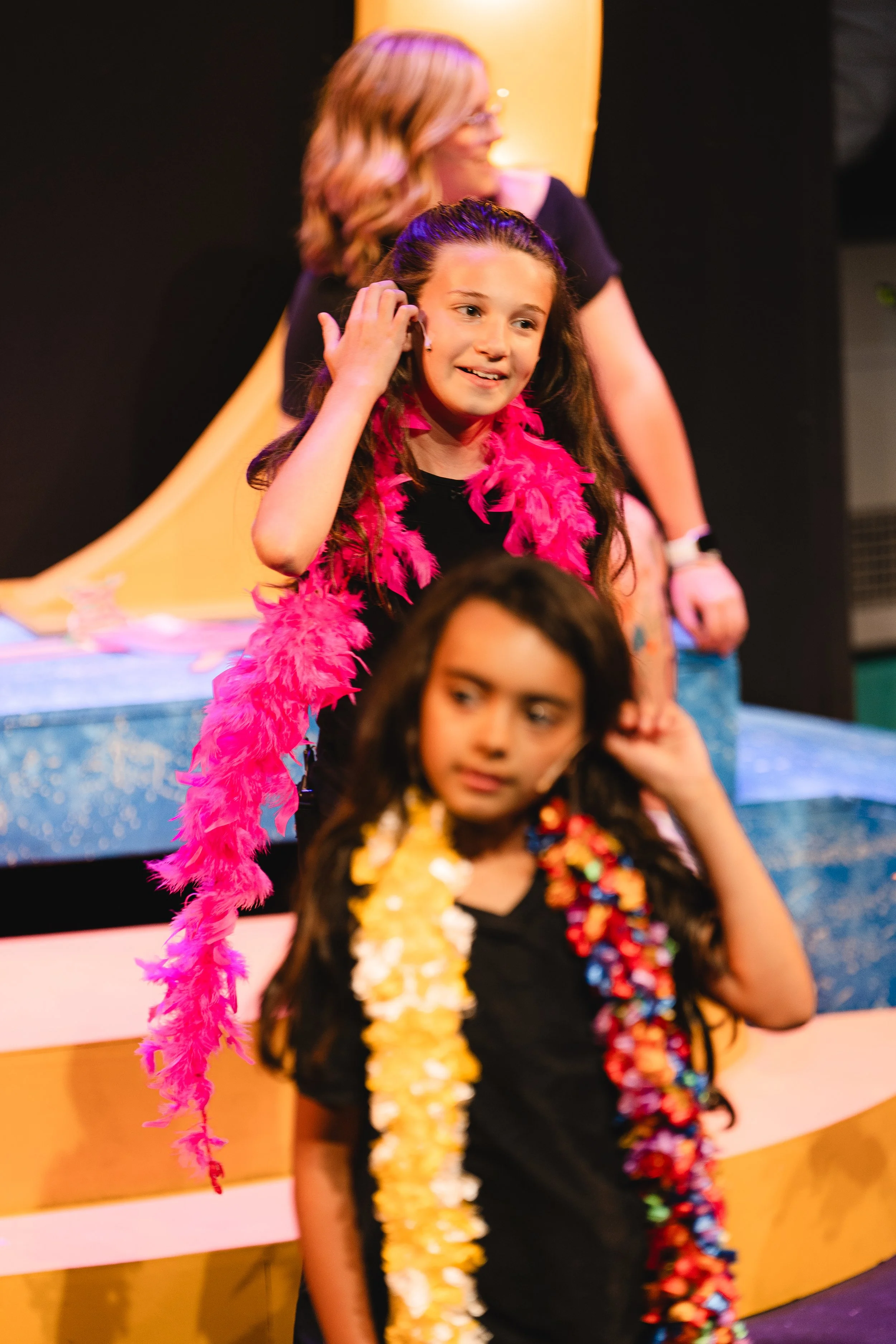 Two young girls dressed in black with colorful leis around their necks, participating in a stage performance or celebration, with one girl adjusting her earring and smiling.