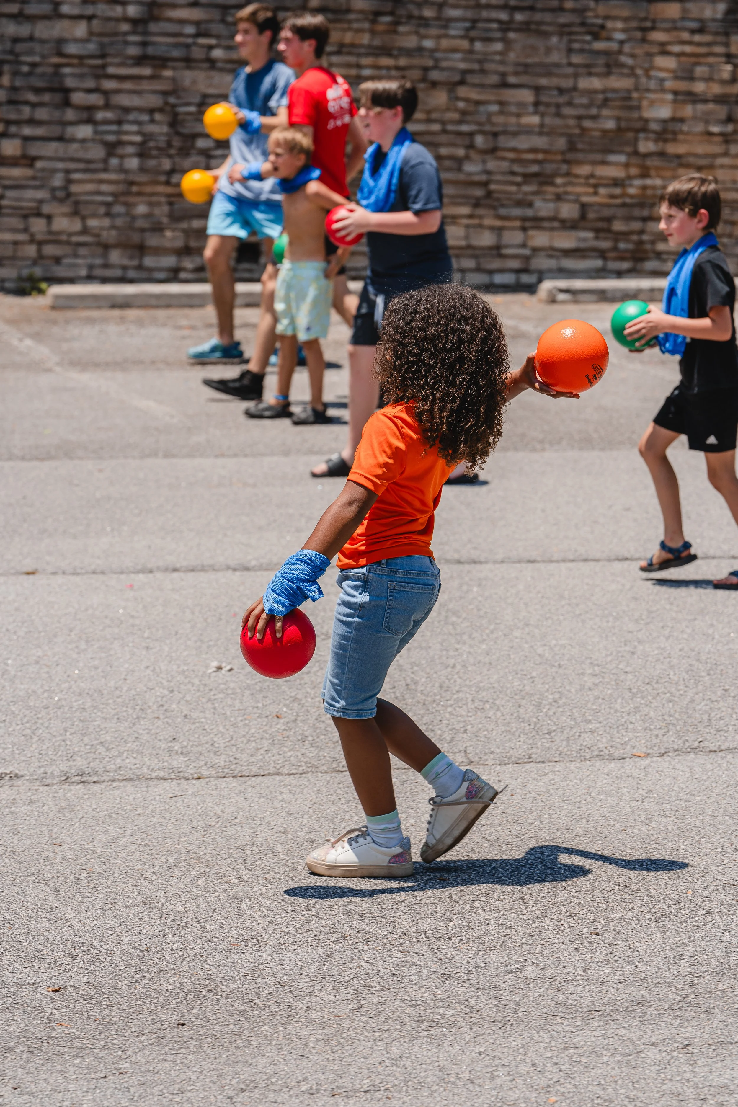 Kids playing with colorful balls outdoors on a sunny day, lined up in a game or activity.