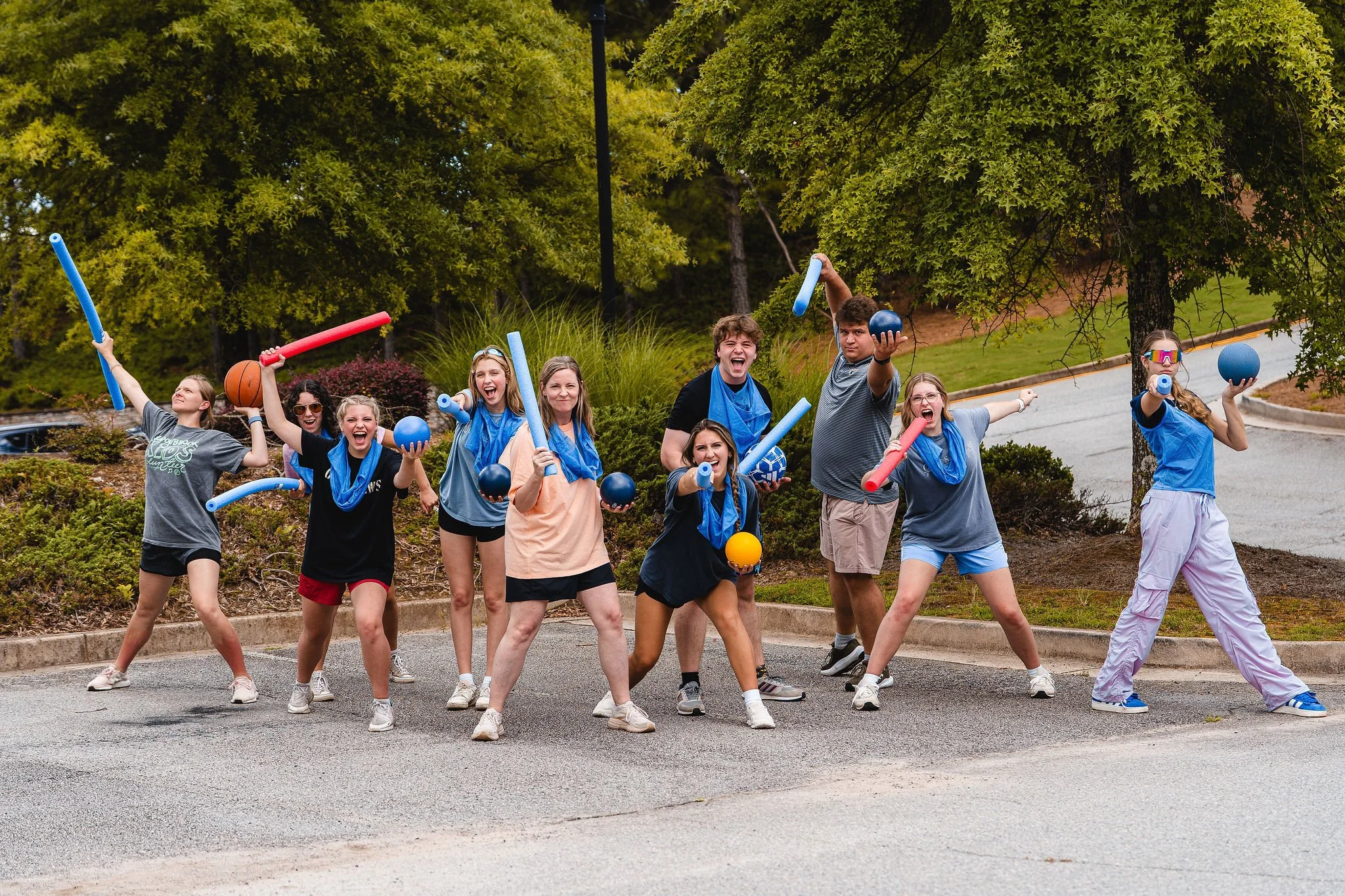Group of children outdoors playing with pool noodles and balls, some wearing blue bandanas, in a park with trees.