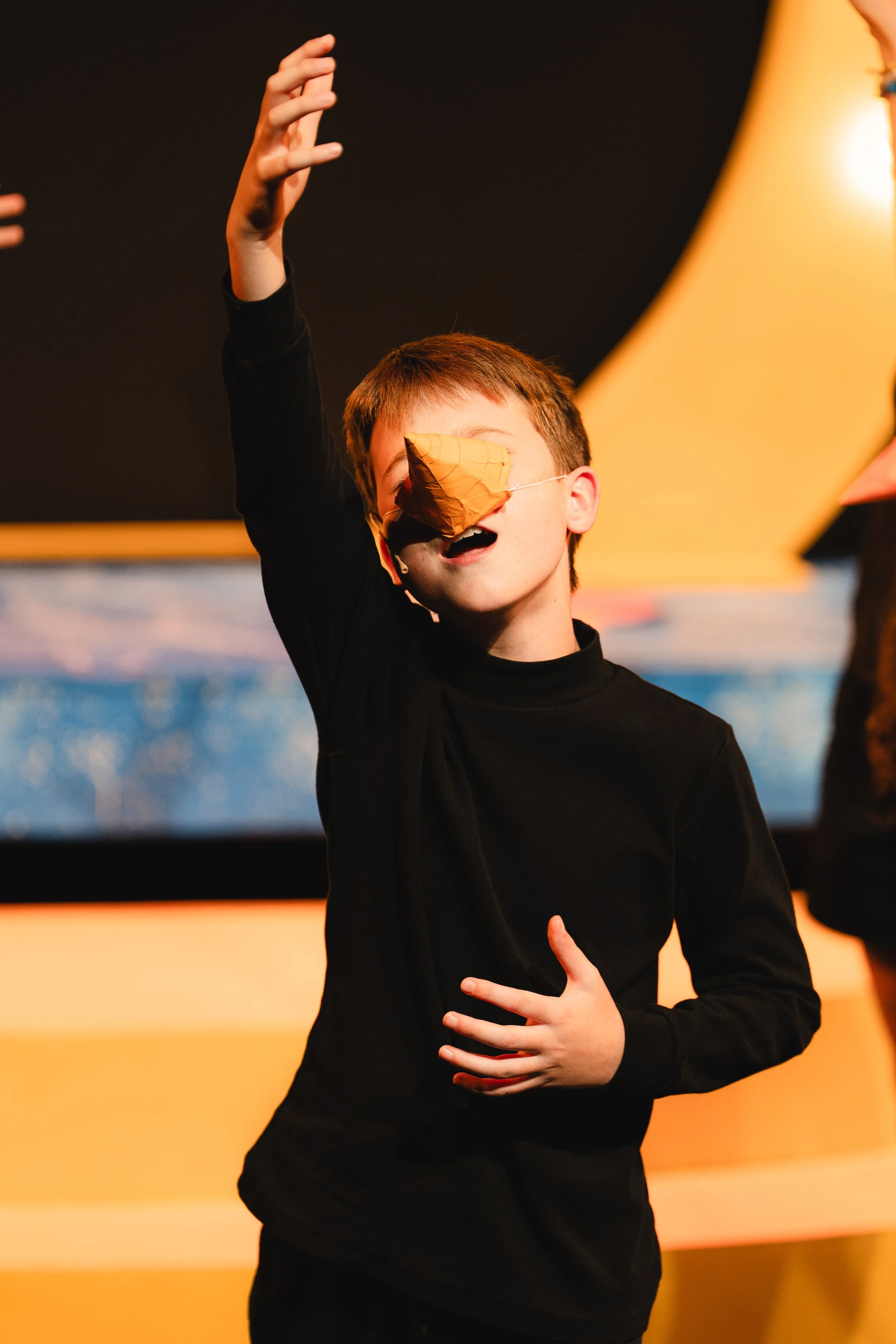 A child with short red hair wearing a black shirt, with a large orange leaf covering their face, hands raised, appearing to be performing on stage with a dark background and warm yellow lighting.