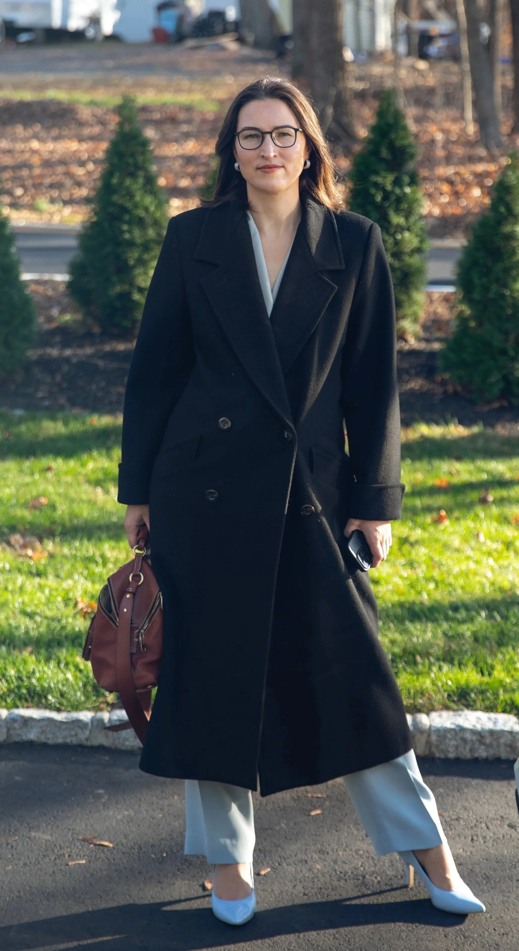 A woman with glasses, wearing a black coat, light-colored pants, and white heels, stands on a paved surface outdoors with green grass and small evergreen trees in the background.