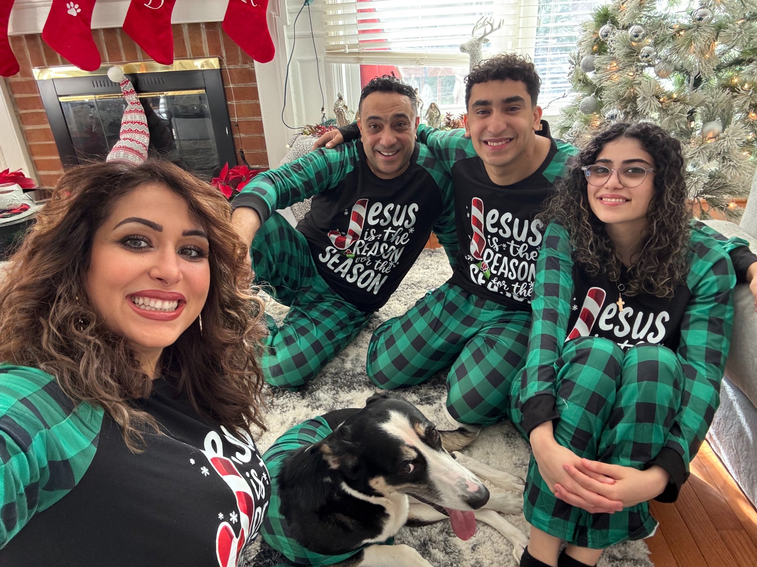 Family of five in matching Christmas pajamas and dog sitting on a carpet next to a Christmas tree, in a living room with stockings hanging on a fireplace.