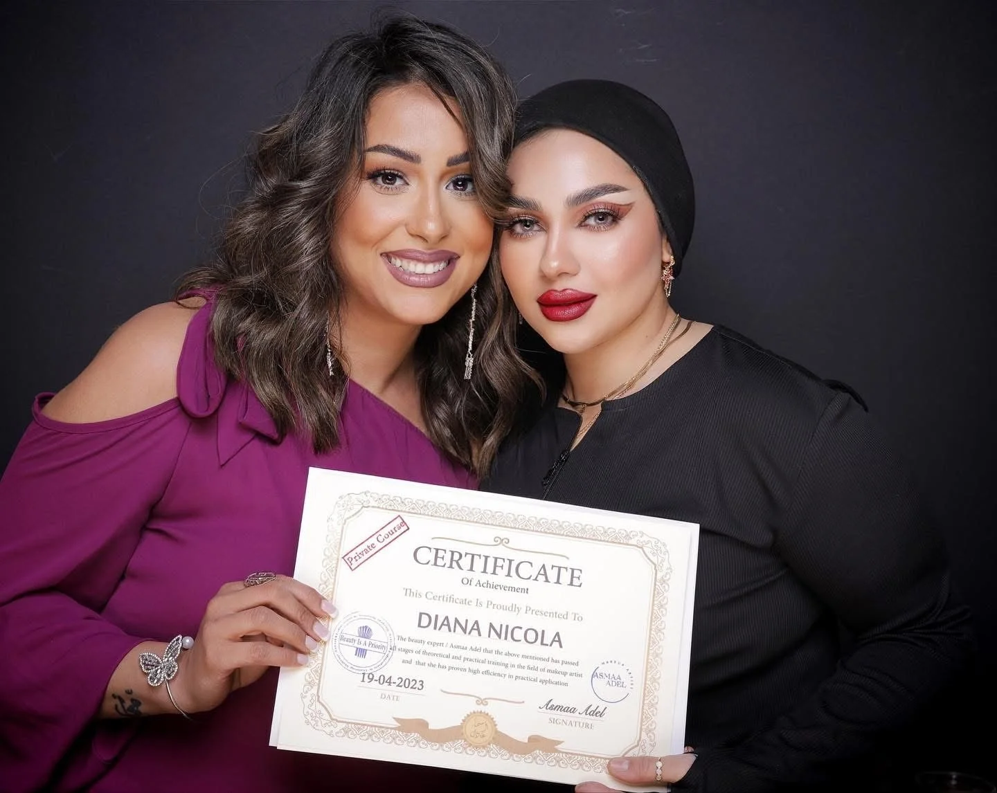 Two women smiling and holding a certificate of achievement. One woman has shoulder-length brown hair and a purple dress, the other has dark hair, red lipstick, and wears a black headscarf and black top.