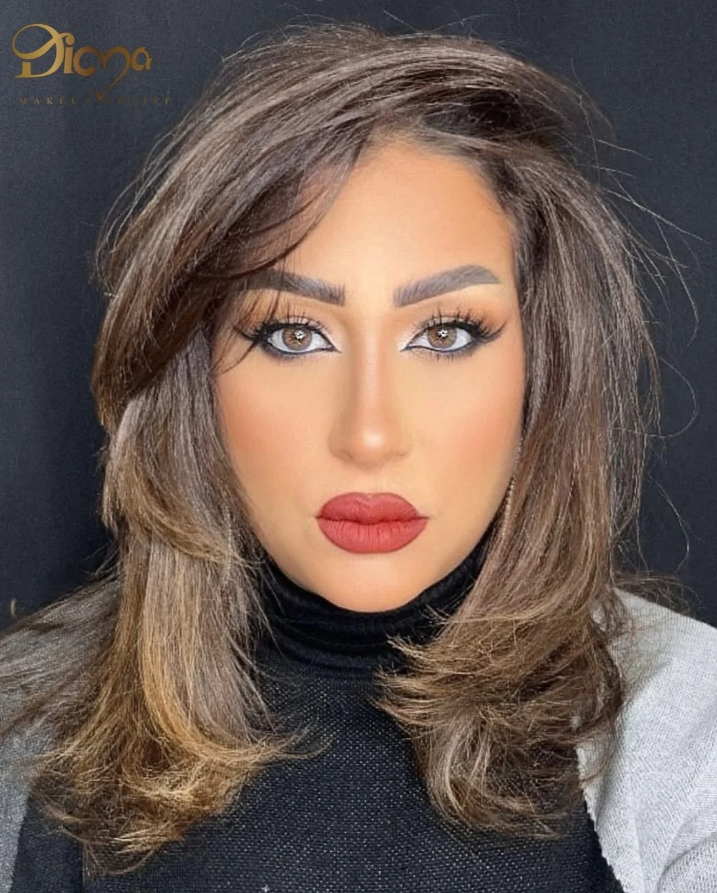 A woman with light brown, wavy hair, light brown eyes, and full glam makeup including red lipstick and defined eyebrows, wearing a black turtleneck and a light-colored blazer. There is a gold logo with the text 'Diana' in the upper left corner.