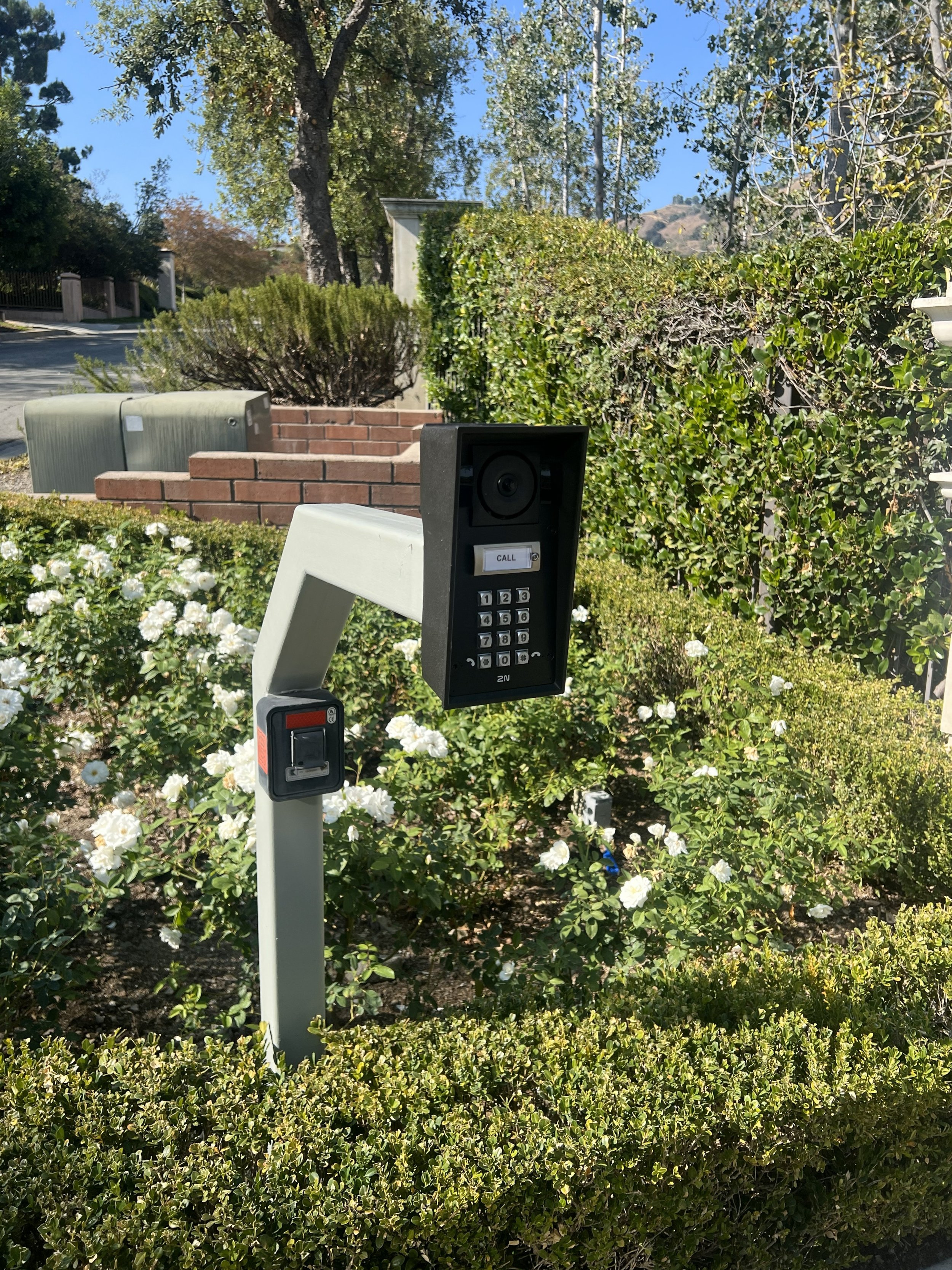 A building intercom system with a camera, call button, keypad, and card reader, installed outdoors among bushes and flowers.