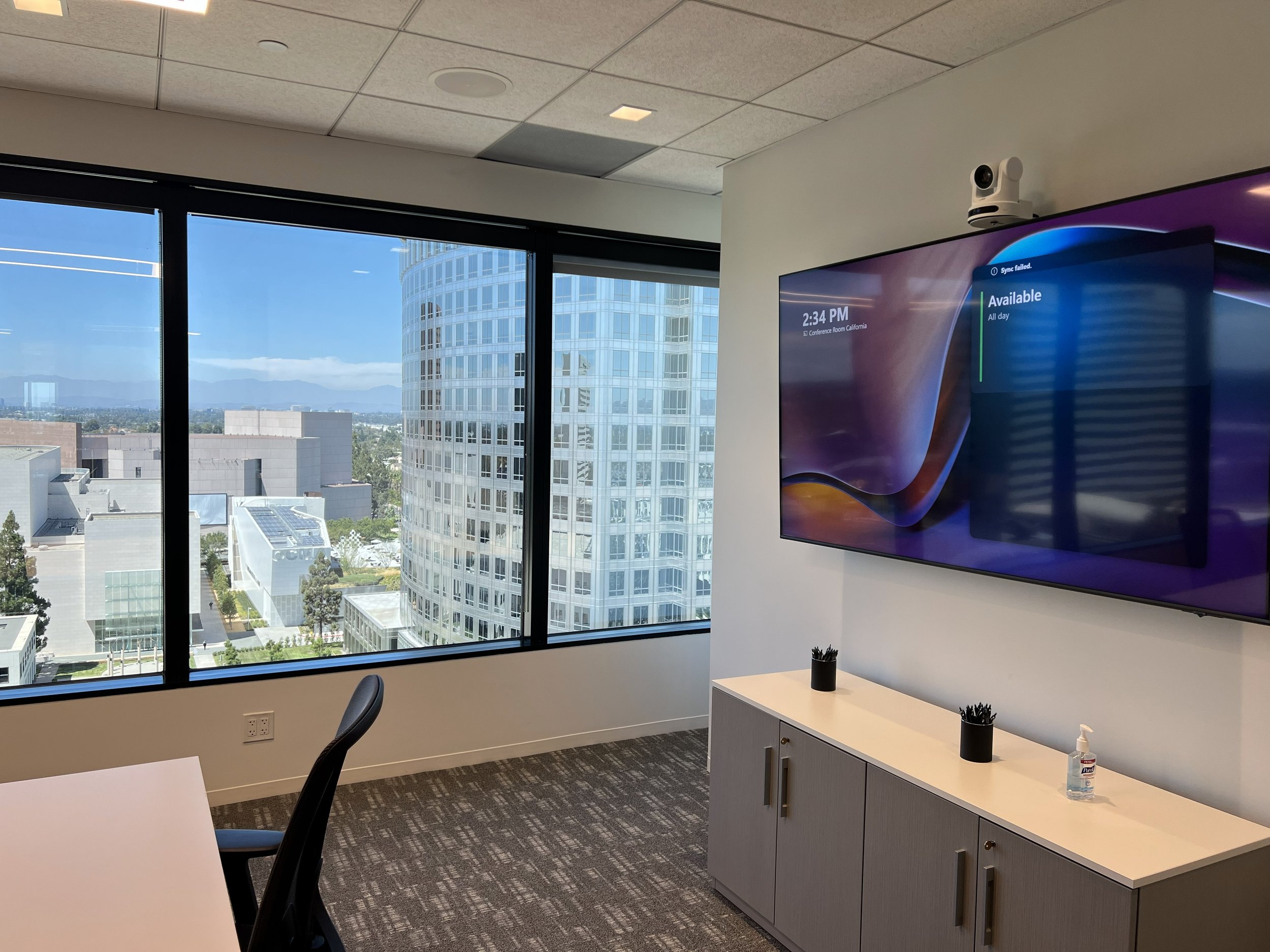 An office conference room with large windows showing a cityscape with tall buildings and mountains in the background. Inside, there is a mounted flat-screen TV on the wall, a cabinet below it with two black containers holding pens and a bottle of hand sanitizer.