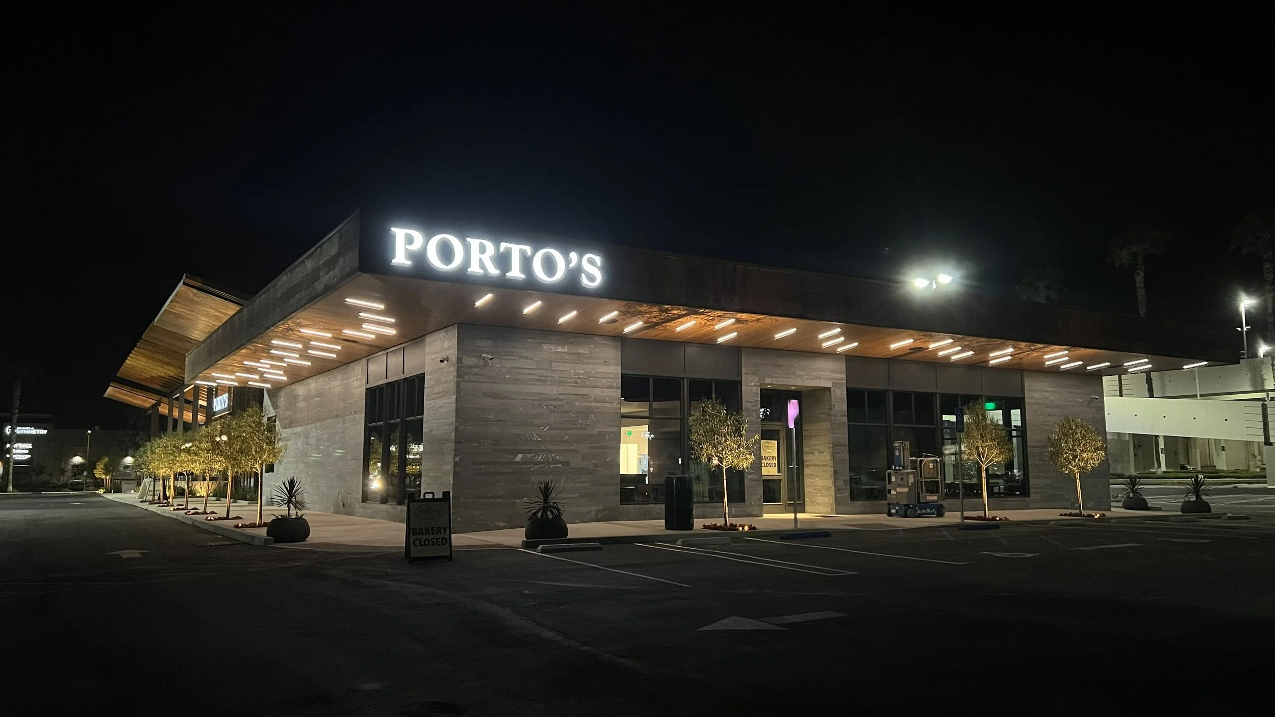 Night view of a modern, single-story restaurant with a large illuminated sign reading 'PORTO'S'. The building has a slate-gray exterior, large windows, and a wooden overhang with embedded lighting. There are small trees in planters and a few parking spaces in front, with one sign indicating the bakery is closed.