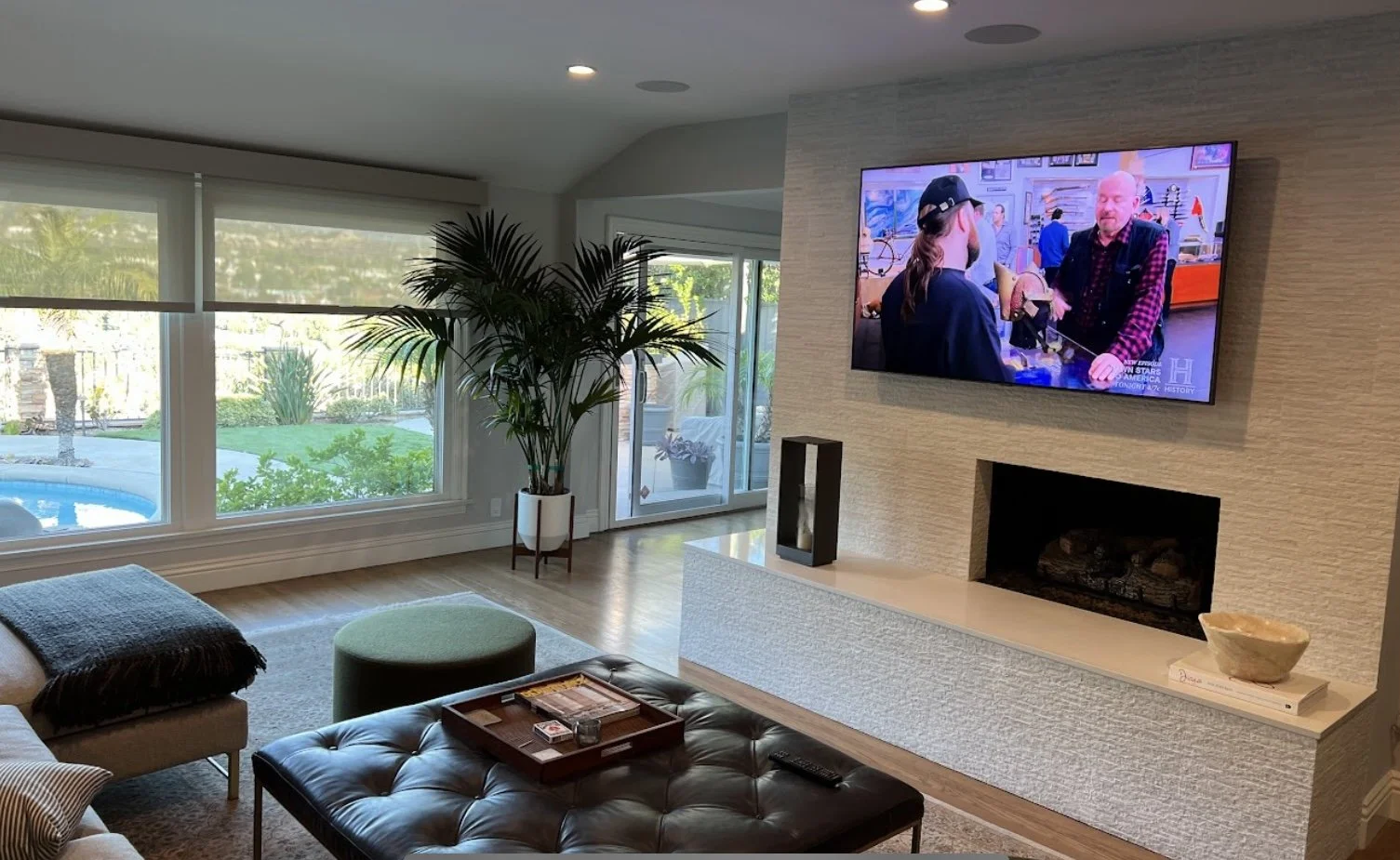 Living room with large windows showing outdoor pool and greenery, a potted plant near sliding glass door, a wall-mounted TV displaying a show, and a modern fireplace beneath the TV. Furniture includes a brown leather tufted ottoman, a sofa with a blanket, a round green ottoman, and a tray with board game pieces and a remote on the ottoman.