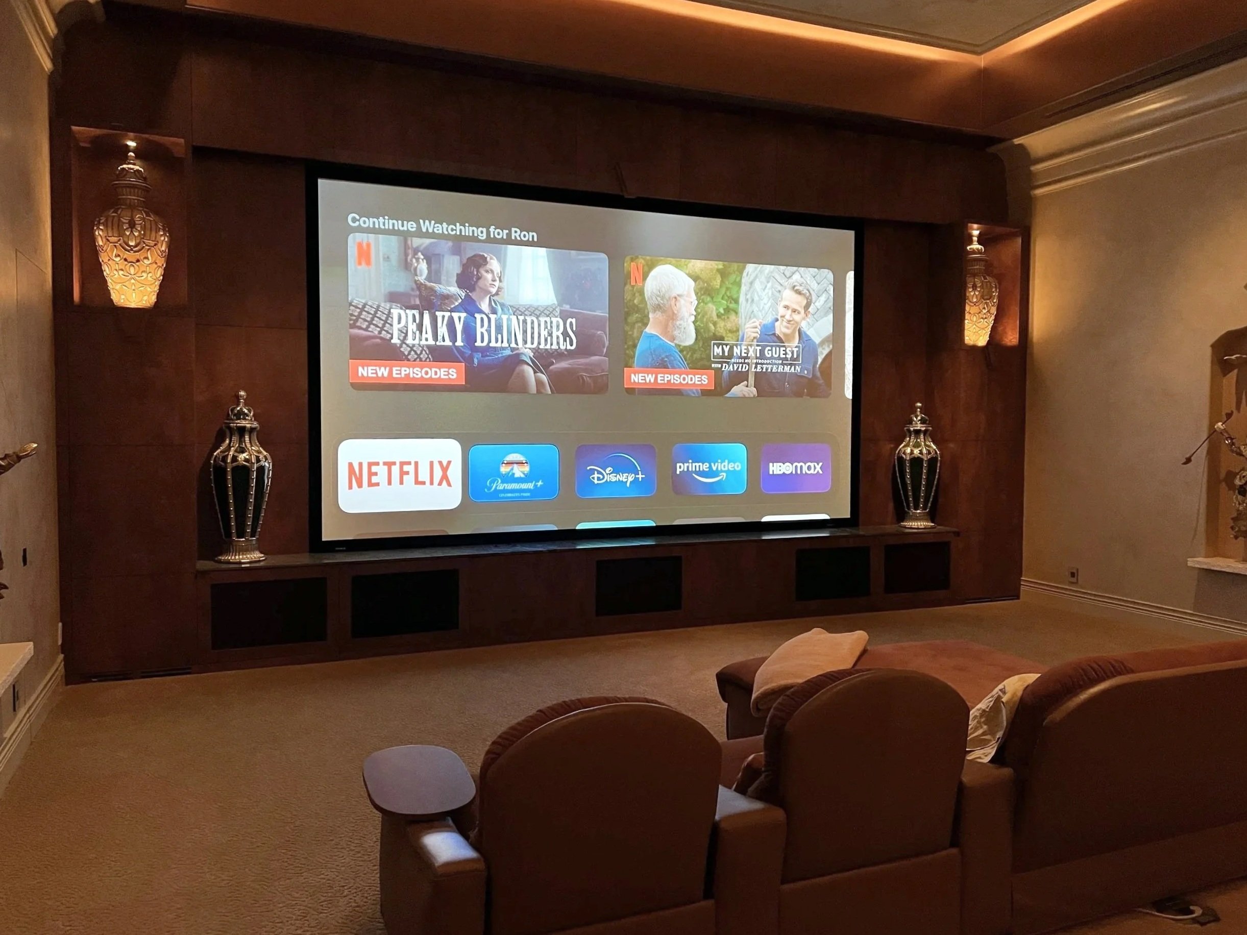 A home theater room with a large screen displaying Netflix's interface, featuring shows 'Peaky Blinders' and 'My Next Guest'. The room has beige carpet, three brown recliner chairs, decorative lamps on either side of the screen, and a small side table.