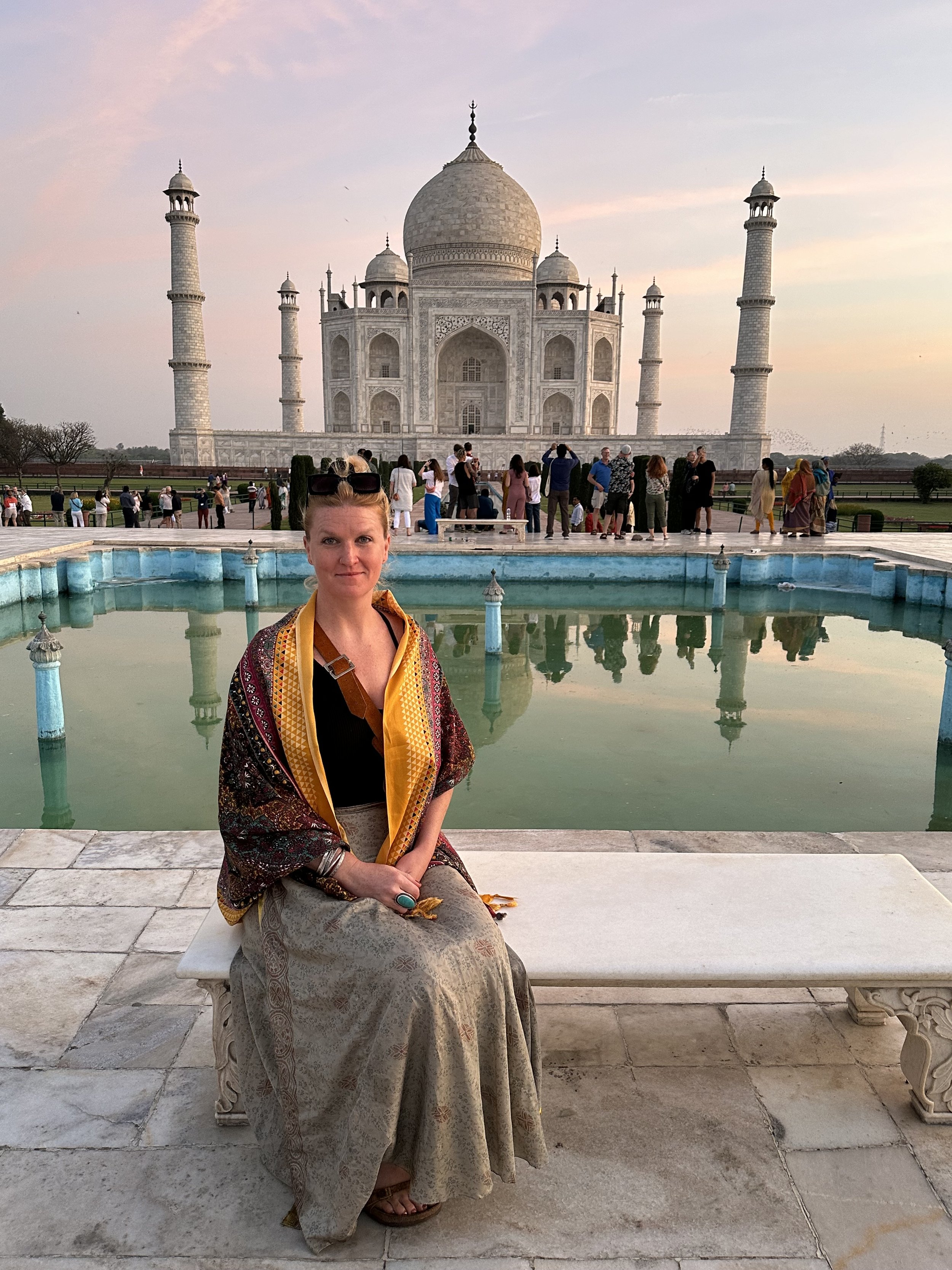 A woman sitting on a white stone bench in front of a reflecting pool, with the Taj Mahal in the background.