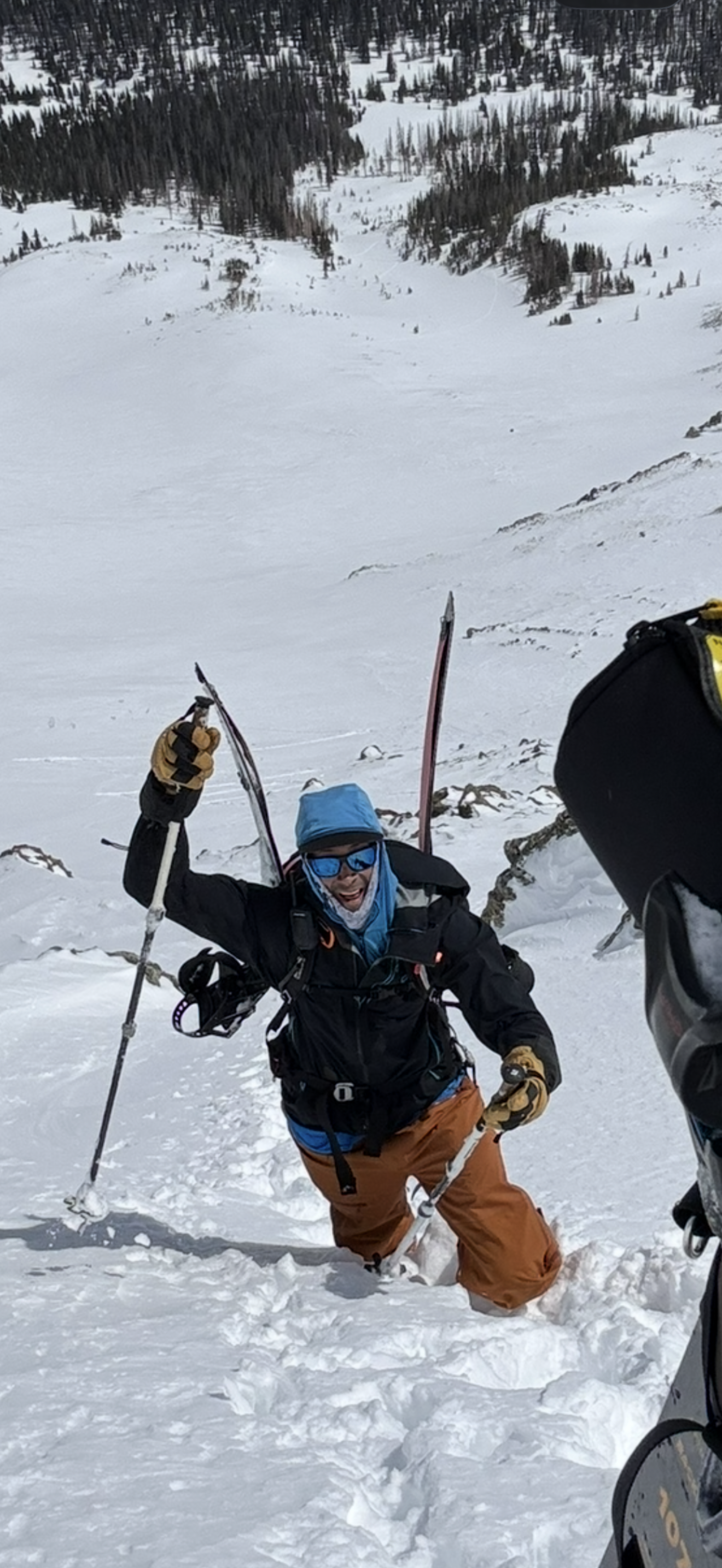 A man dressed in winter gear, wearing sunglasses and gloves, standing in deep snow on a mountain slope, holding ski poles, with a snowy mountain landscape in the background.