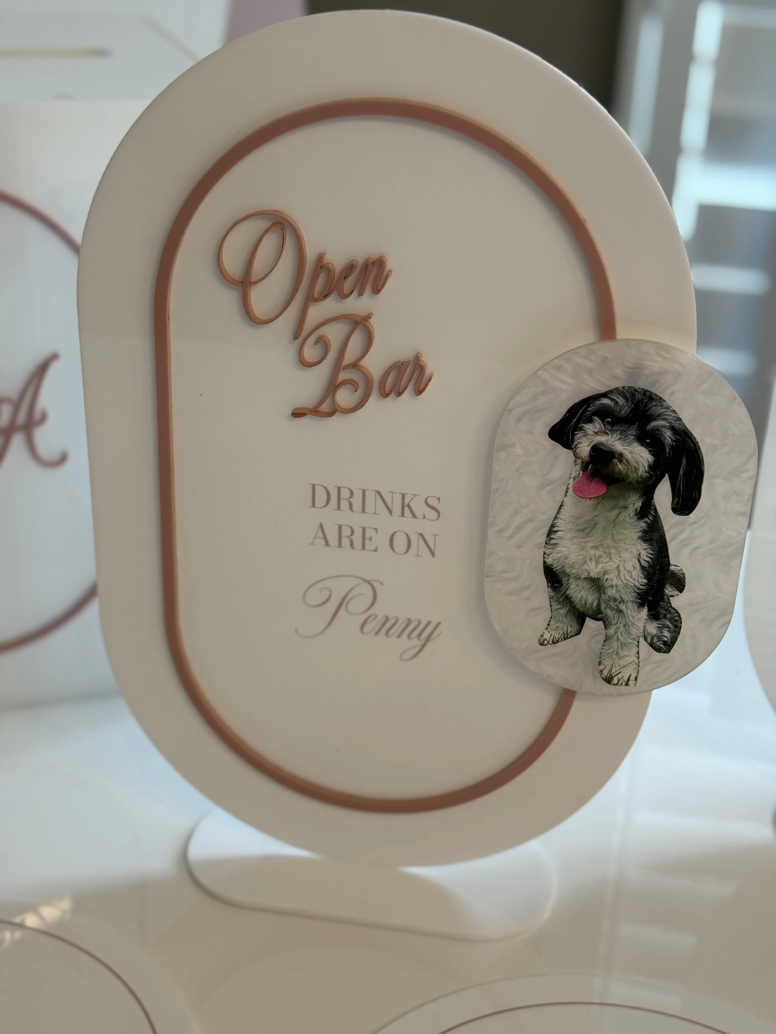 A white sign with pink text says, 'Open Bar' and 'Drinks are on Penny,' with a picture of a cute black and white puppy with its tongue out attached to the sign.