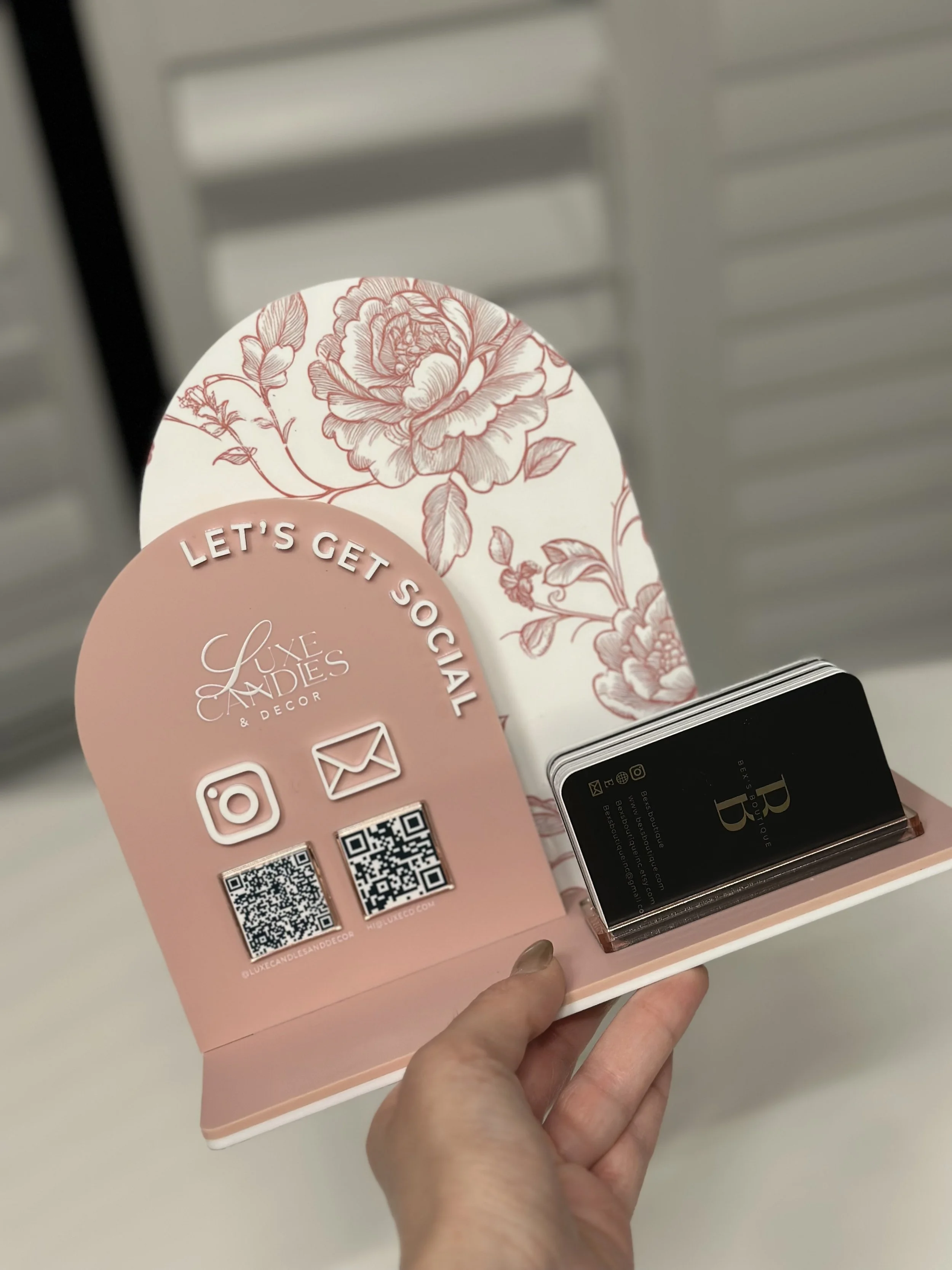 Pink and white floral business card holder with social media icons and QR codes for Luxe Candies & Decor, featuring a black card with a gold letter 'B' and contact info, and pink floral background.