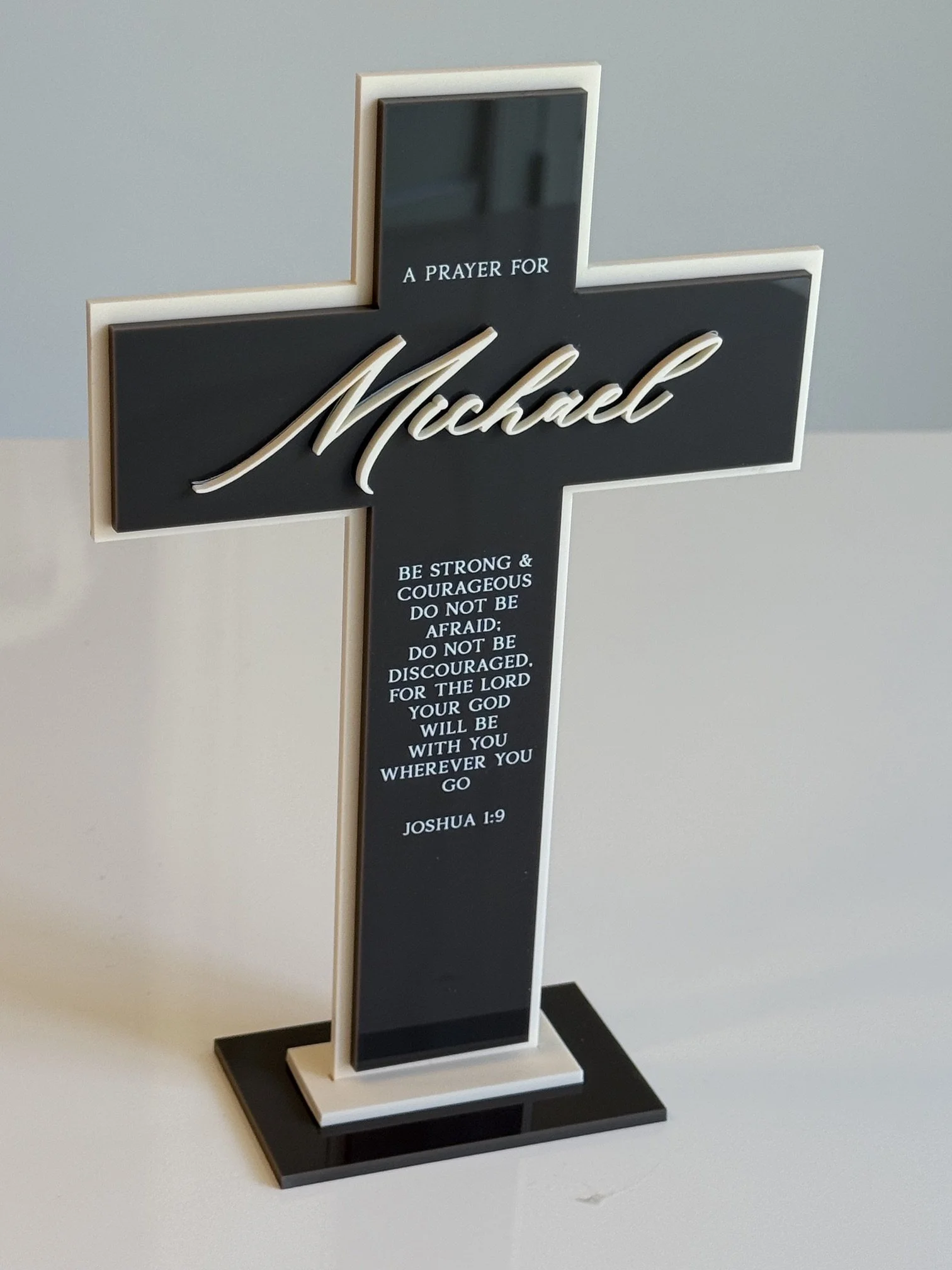 A decorative black and white cross with a scripture quote from Joshua 1:9, reading 'Be strong and courageous. Do not be afraid; do not be discouraged. For the Lord your God will be with you wherever you go.'
