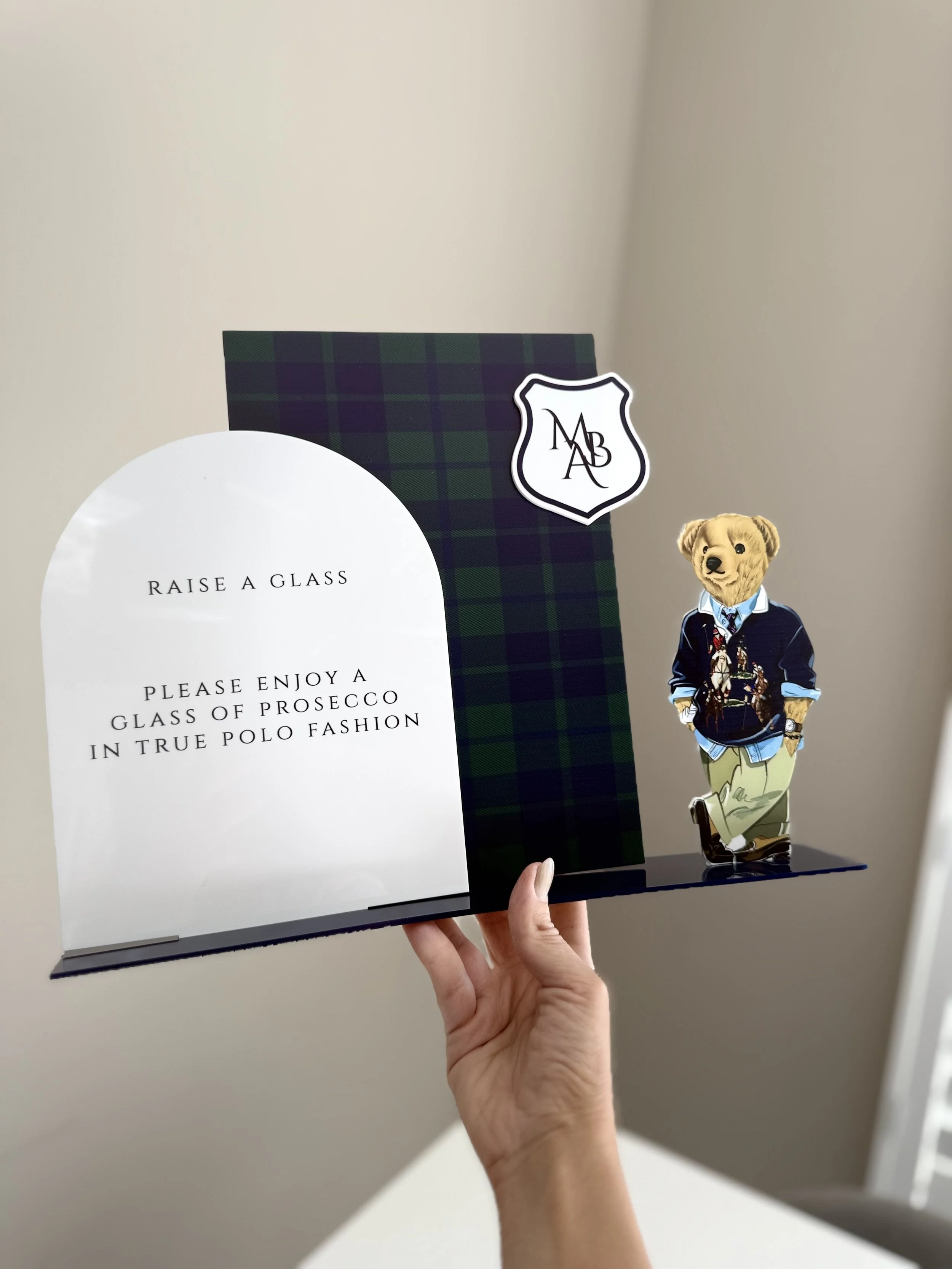 A hand holding a display featuring a white sign with black text, a green and navy plaid background with a logo, and a cutout of a bear dressed in a blue jacket and light pants.