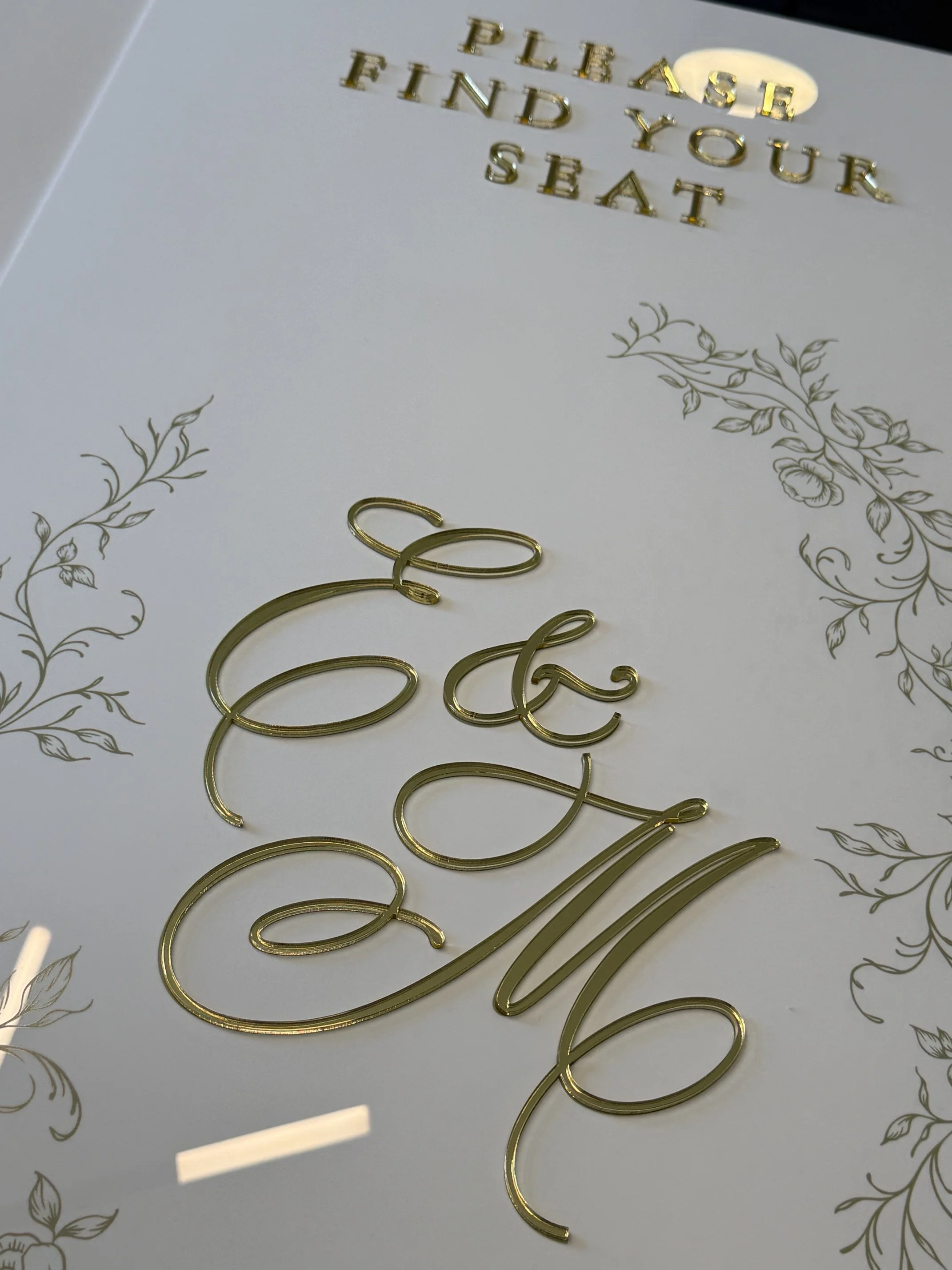 Close-up of a greeting card with gold embossed words "Please find your seat" and decorative floral designs, with gold cursive lettering.