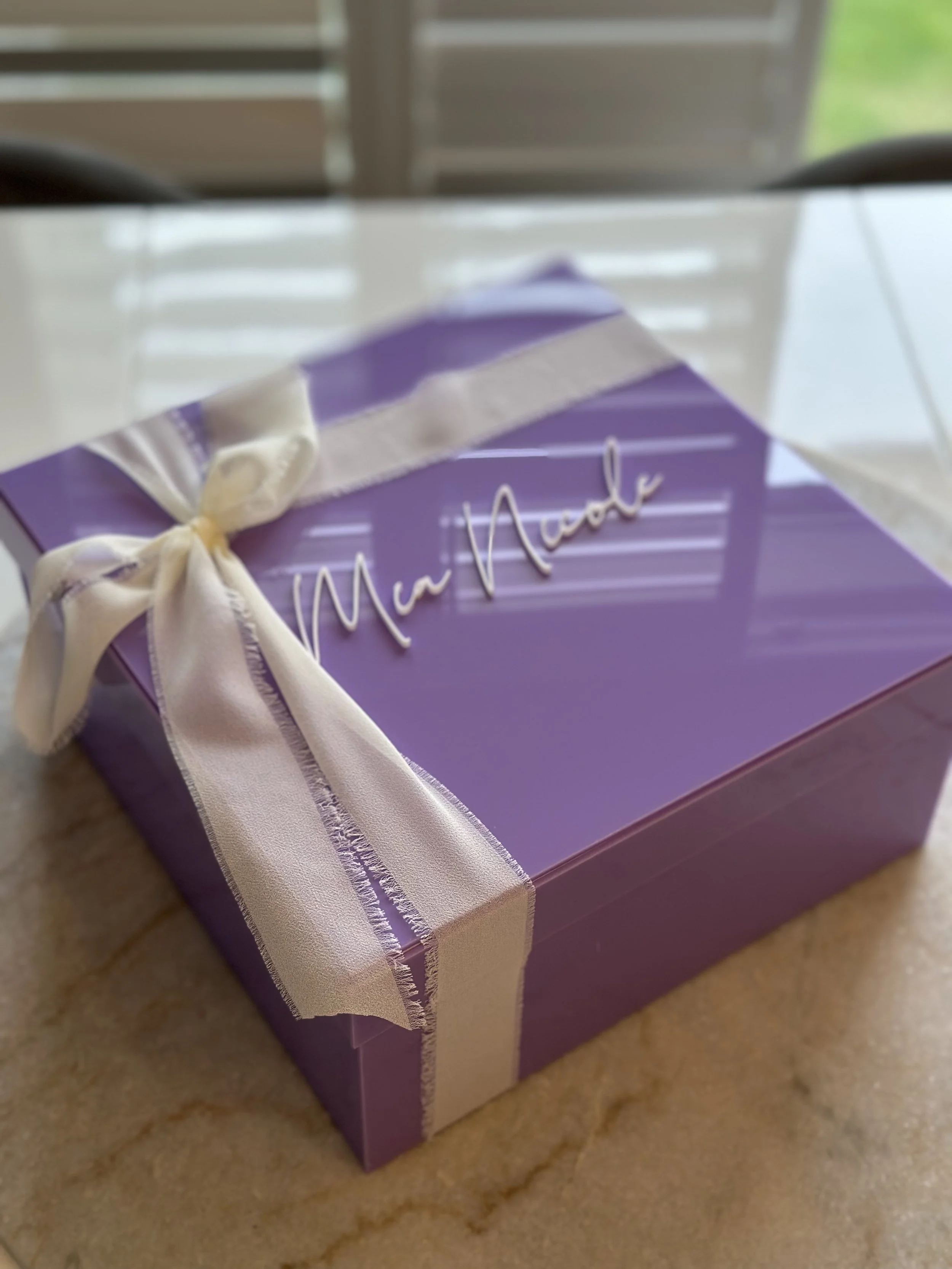 Purple gift box with a white ribbon and bow, with 'Mu Mu Naomi' written on the top, placed on a light-colored surface.