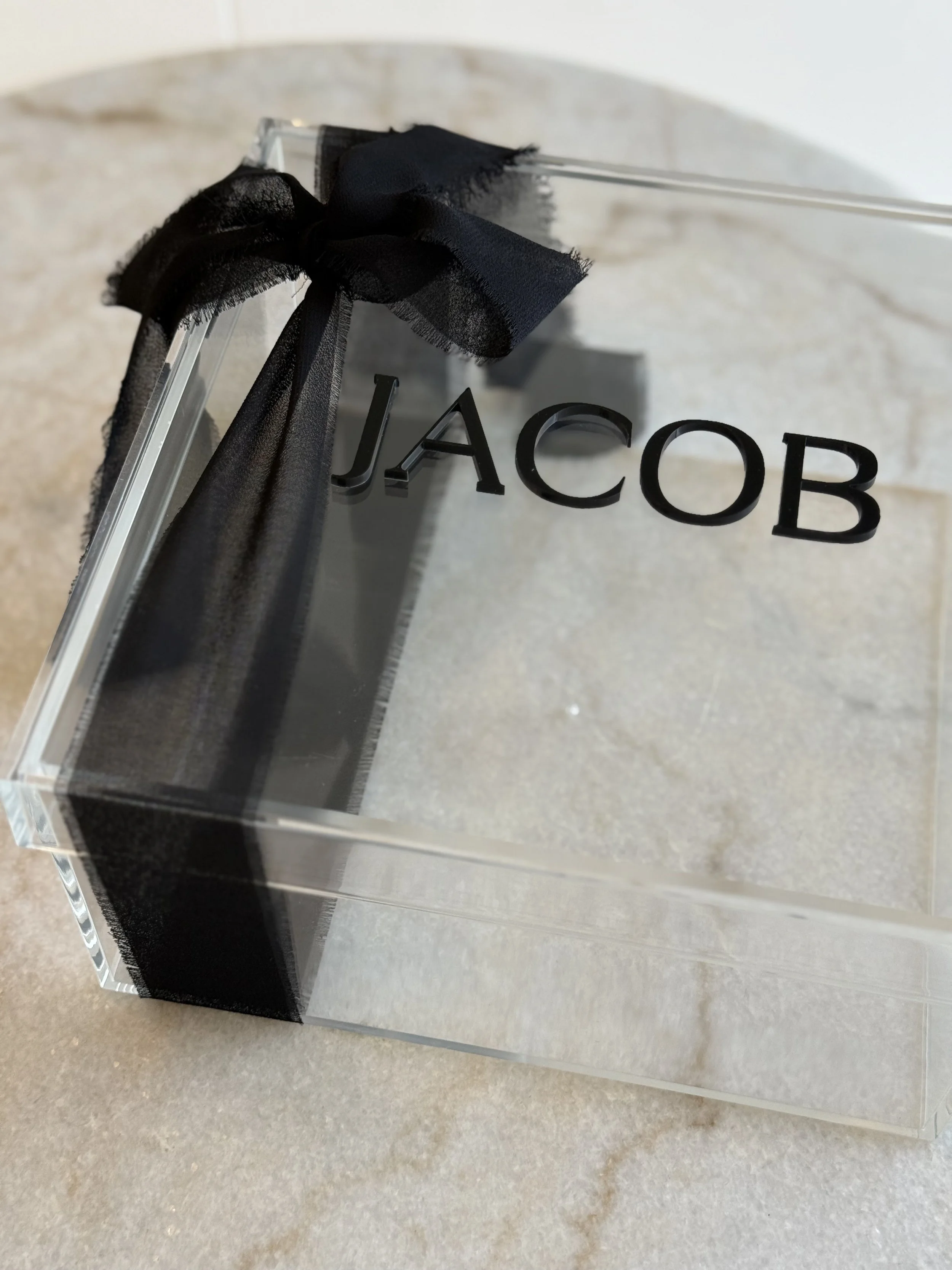 A clear acrylic box with the name 'JACOB' in black 3D letters, decorated with a black ribbon tied into a bow on one corner, sitting on a light-colored surface.