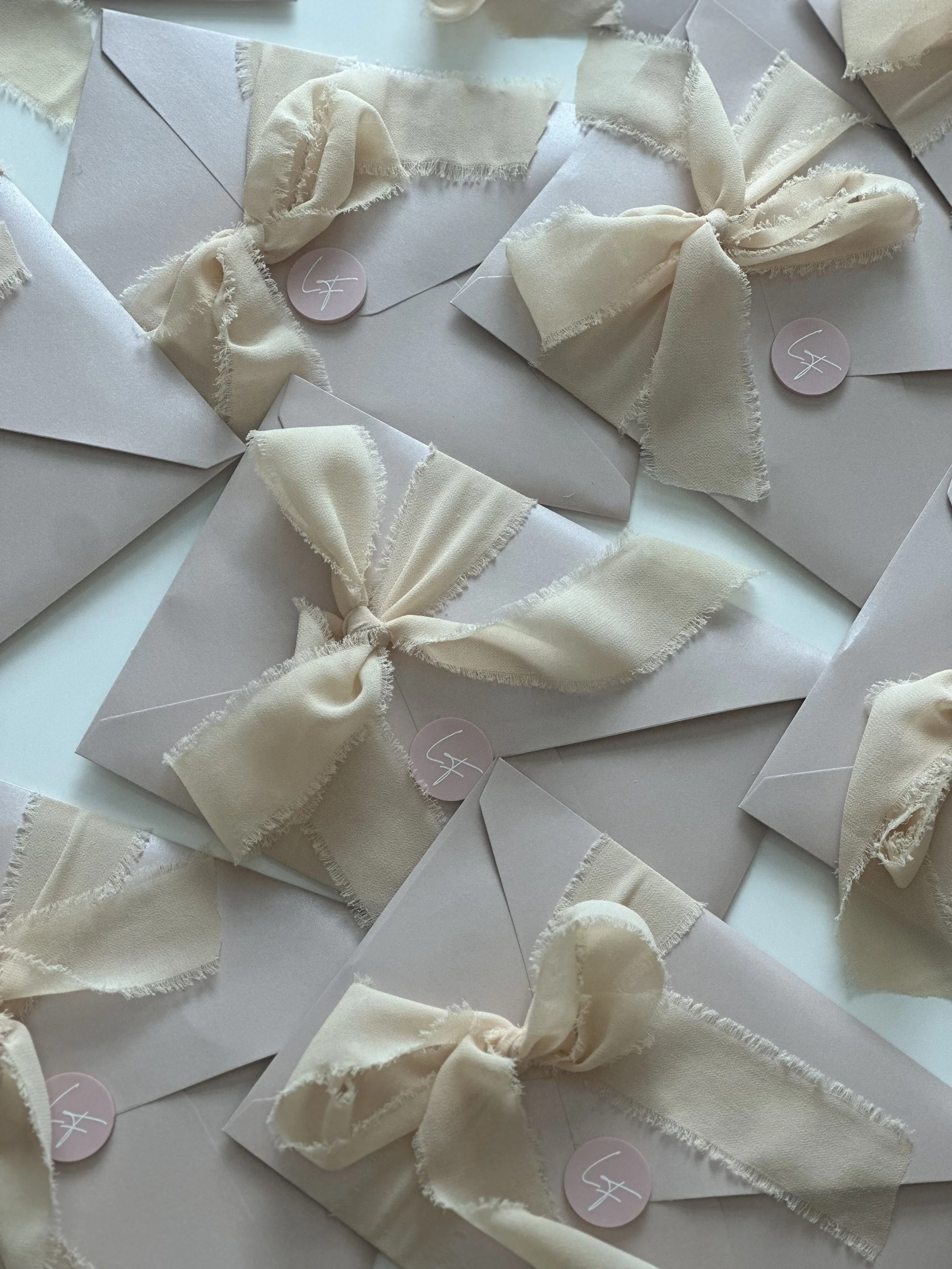Multiple white envelopes decorated with cream-colored fabric bows attached to pink circular stickers with a white silhouette of an 'F'.