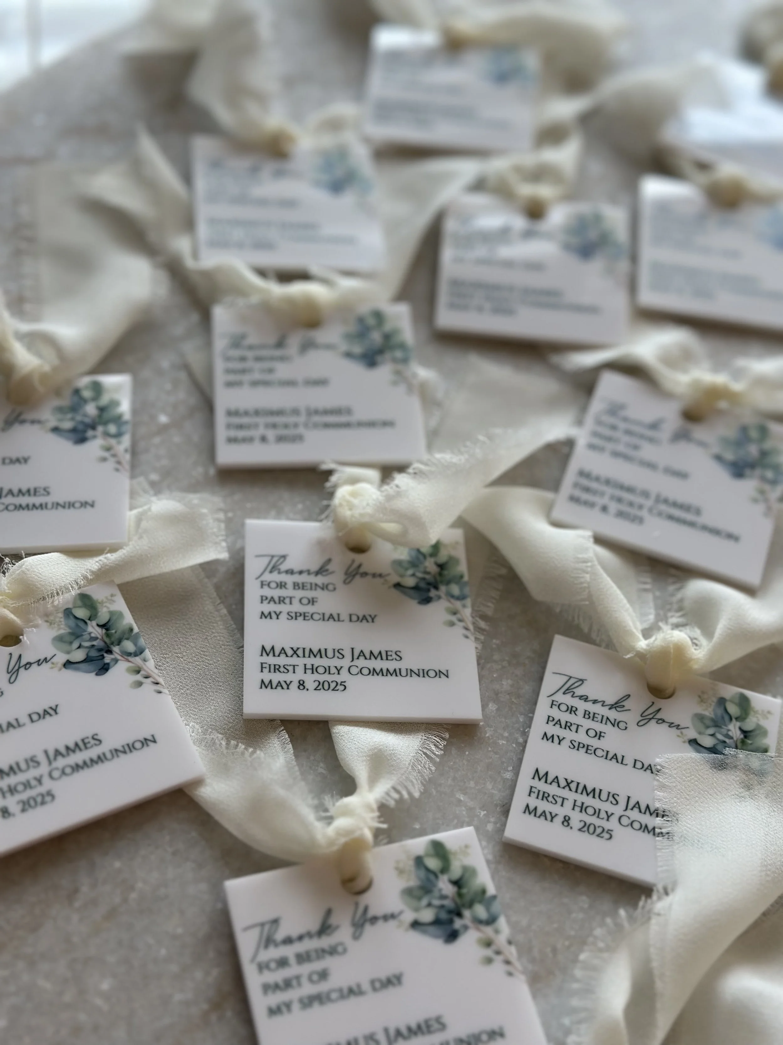 Multiple white cards with three small green plants artwork and black text, attached to white fabric ribbons, on a table. The cards thank guests for being part of a special day and indicate a First Holy Communion on May 8, 2025.