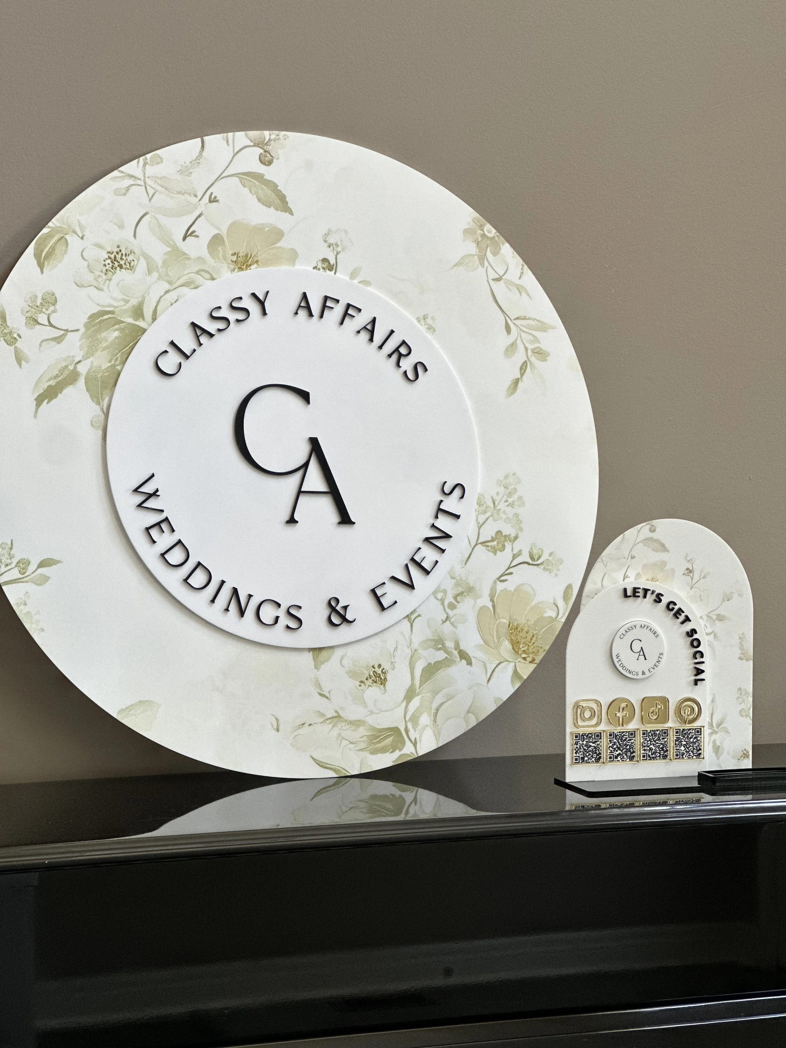 Decorative sign for Classy Affairs Weddings & Events, featuring a floral background with the company logo and social media QR codes, placed on a countertop.