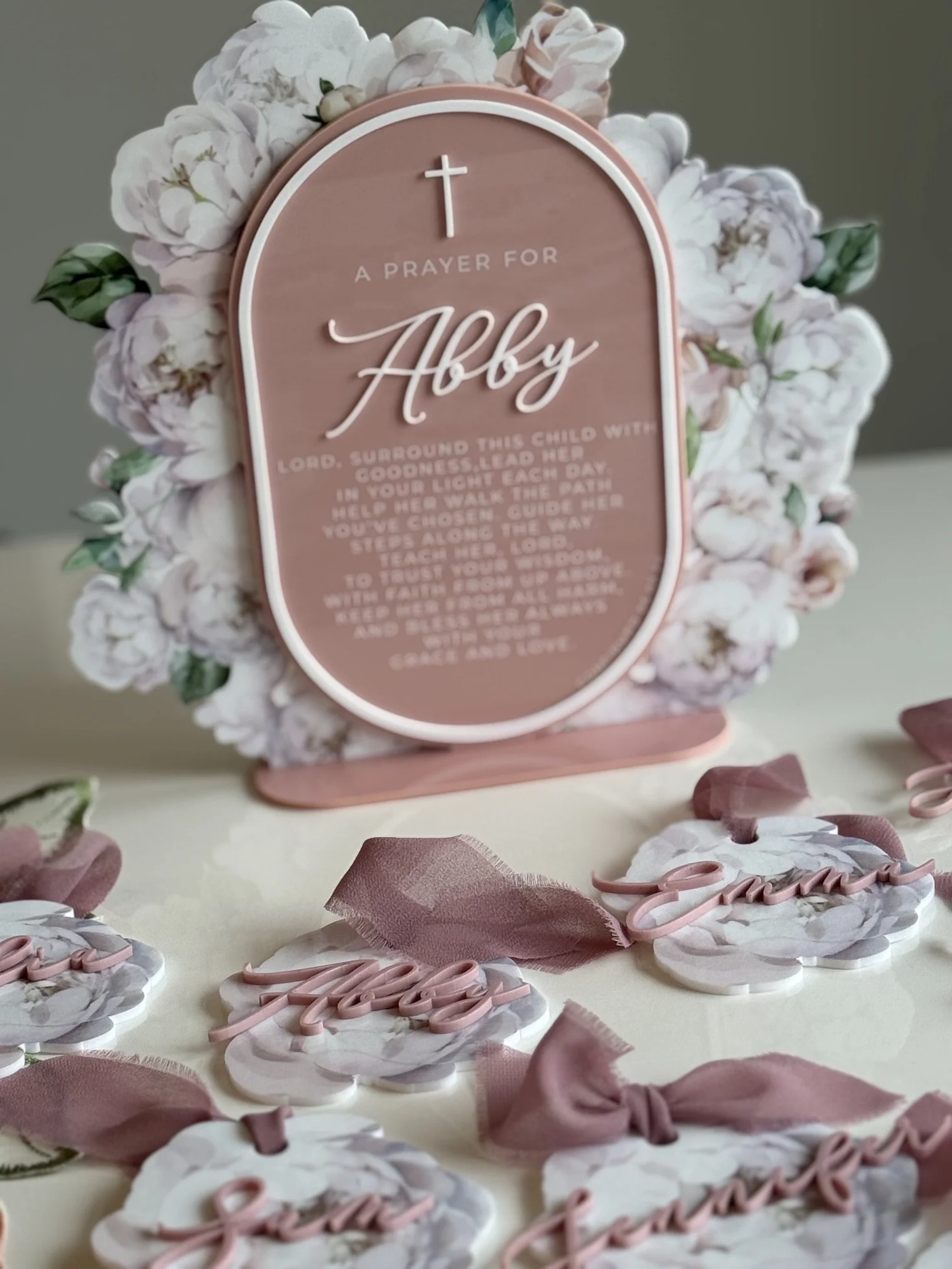 A pink plaque with a white cross at the top and the words 'A Prayer for Abigail' in cursive, surrounded by white flower decorations, on a table scattered with pink and white flower-shaped cookies and pink ribbon bows.