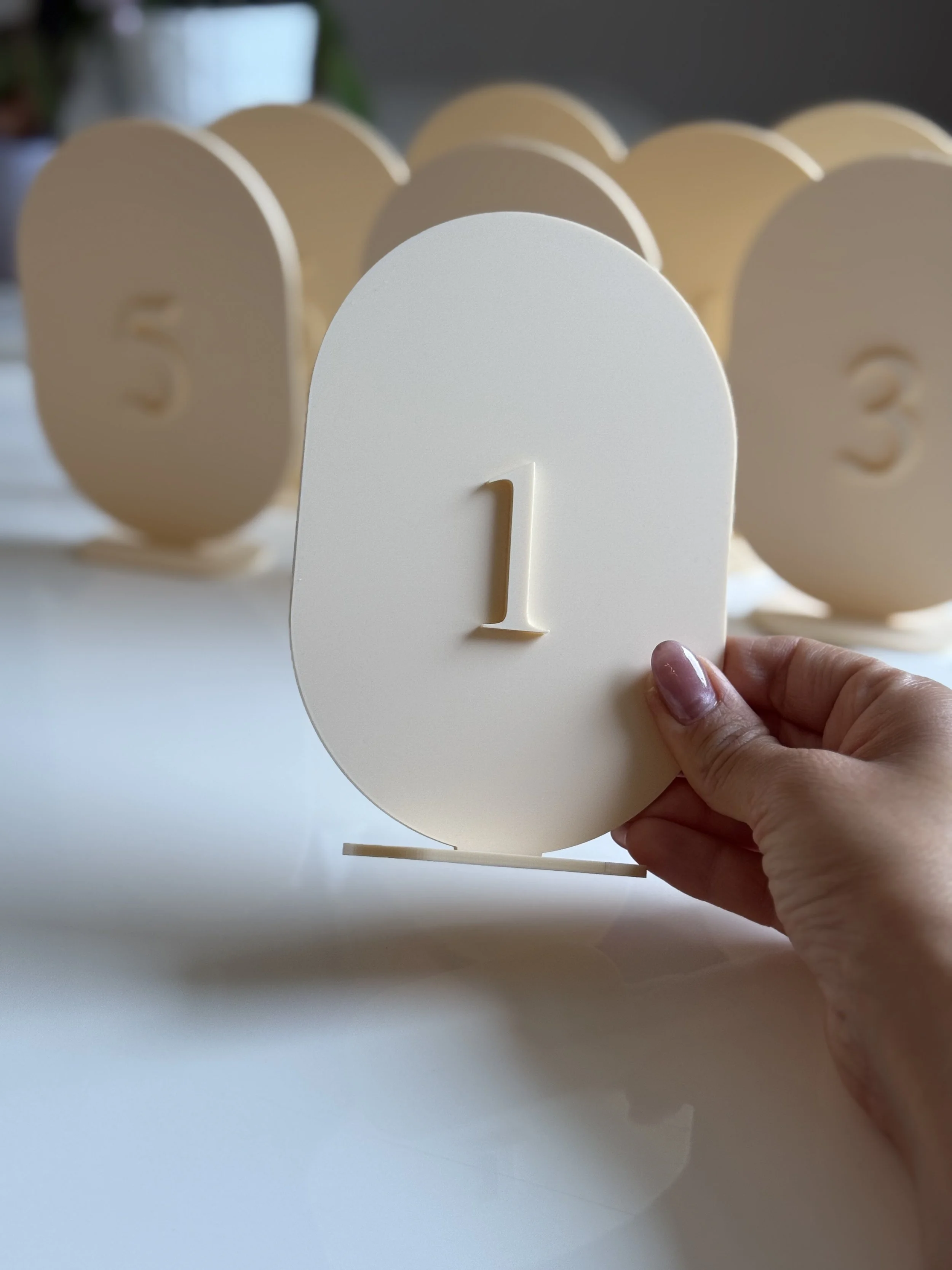 A hand holding a cream-colored stand with the number 1 in a raised design, with other similar stands with numbers 2, 3, 4, and 5 in the background.