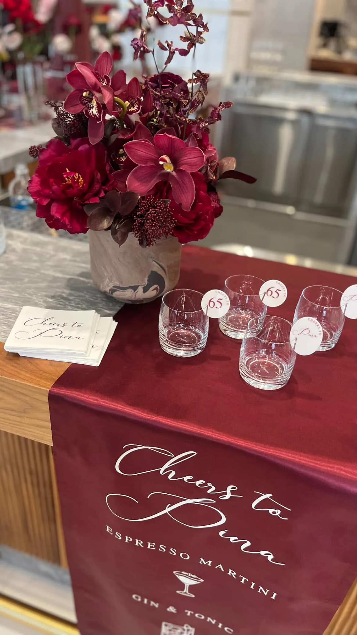 A floral arrangement with burgundy, pink, and purple flowers in a marble vase, placed on a table with a burgundy tablecloth that reads 'Cheers to Piña' and 'Espresso Martini.' Small glasses with tags marked '65' are arranged beside the flowers. There