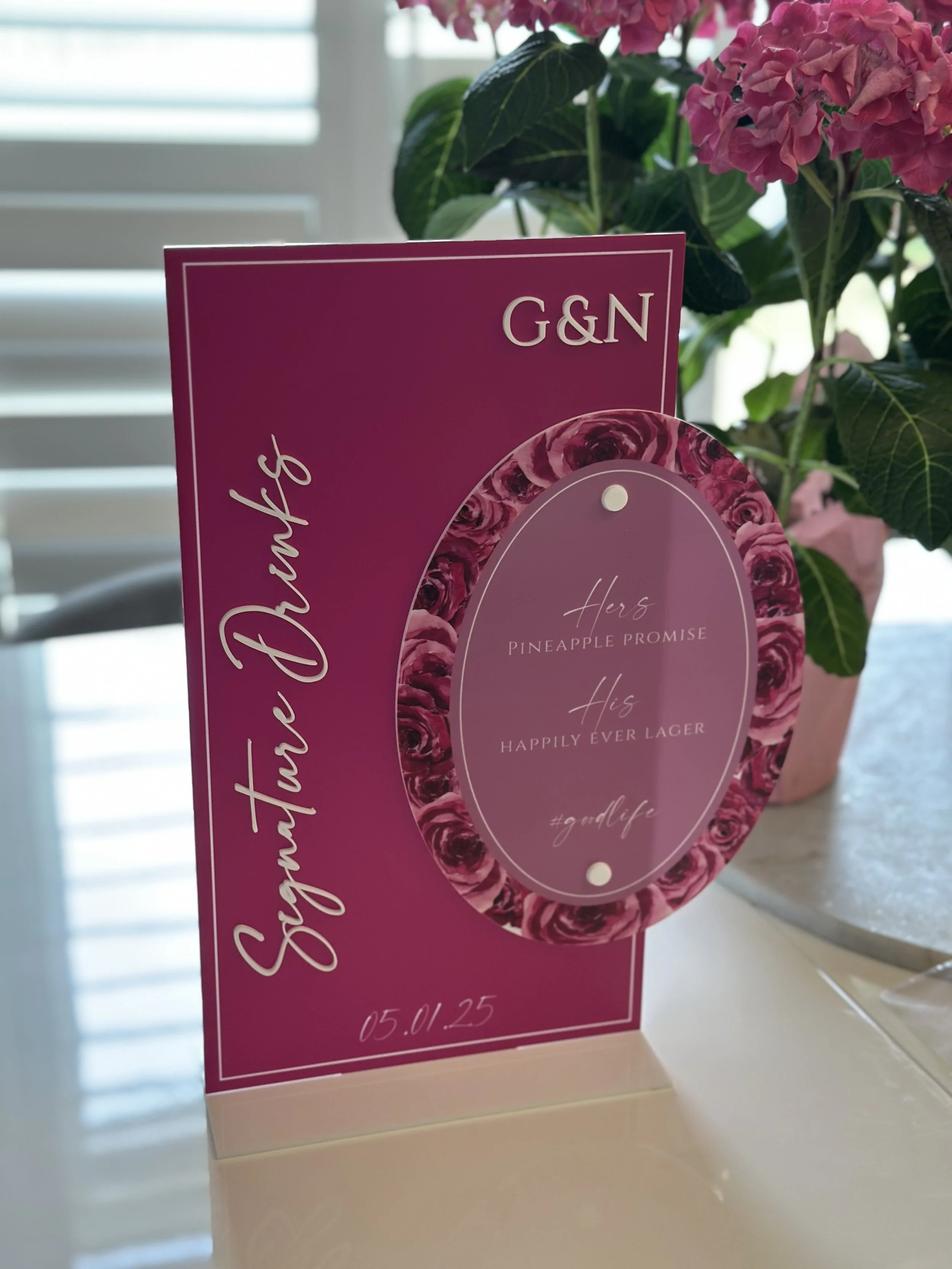 A pink card with floral design elements reading 'Sergio D'angelo G&N' and '05.01.25'; a circular element with a rose border that says 'Here's Pineapple Promise. His Happily Ever Lager #gudelife'; pink hydrangea flowers in a pot in the background.