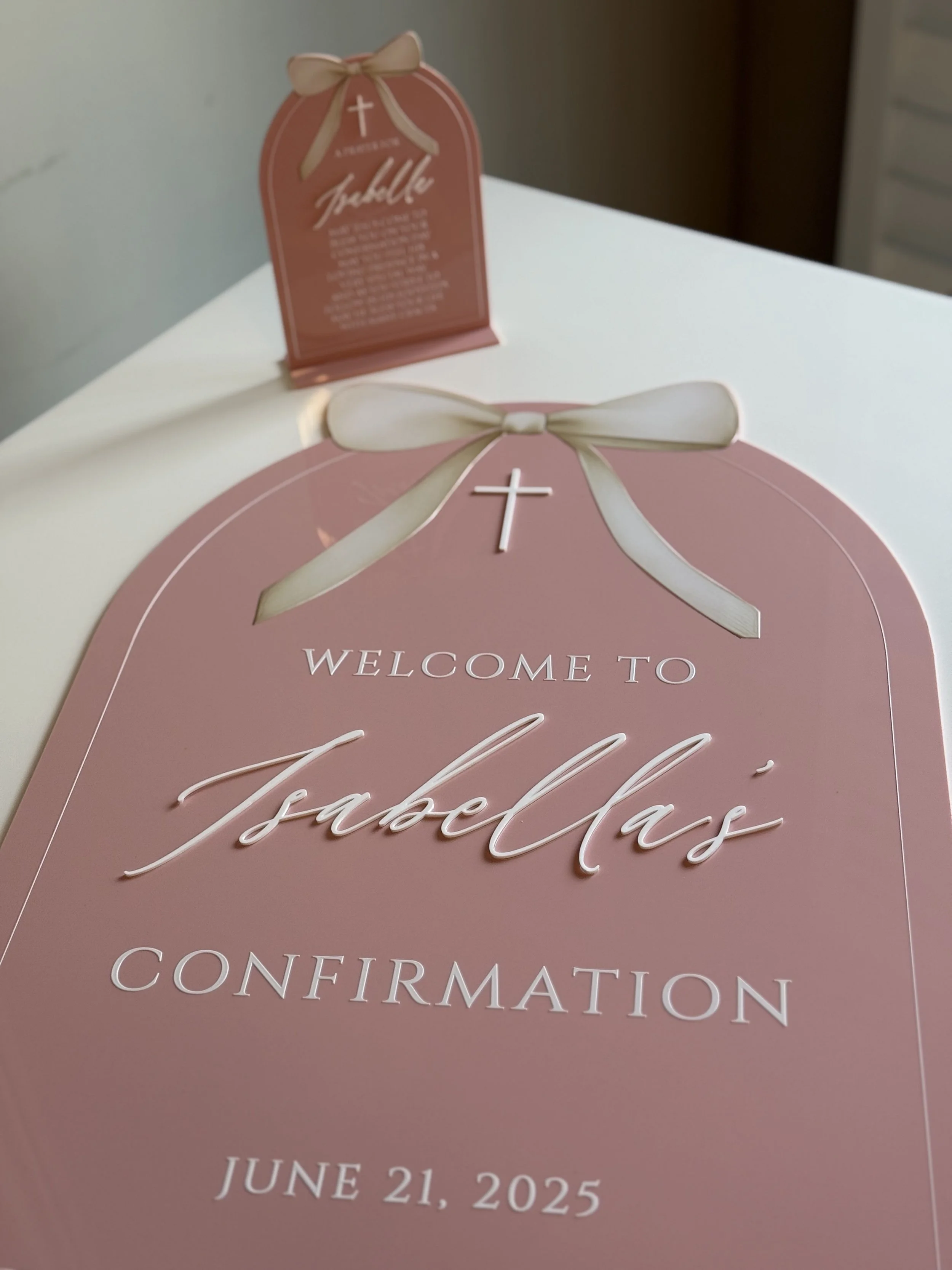 Pink and white welcome sign for Isabel's confirmation on June 21, 2025, decorated with a bow and a small cross.