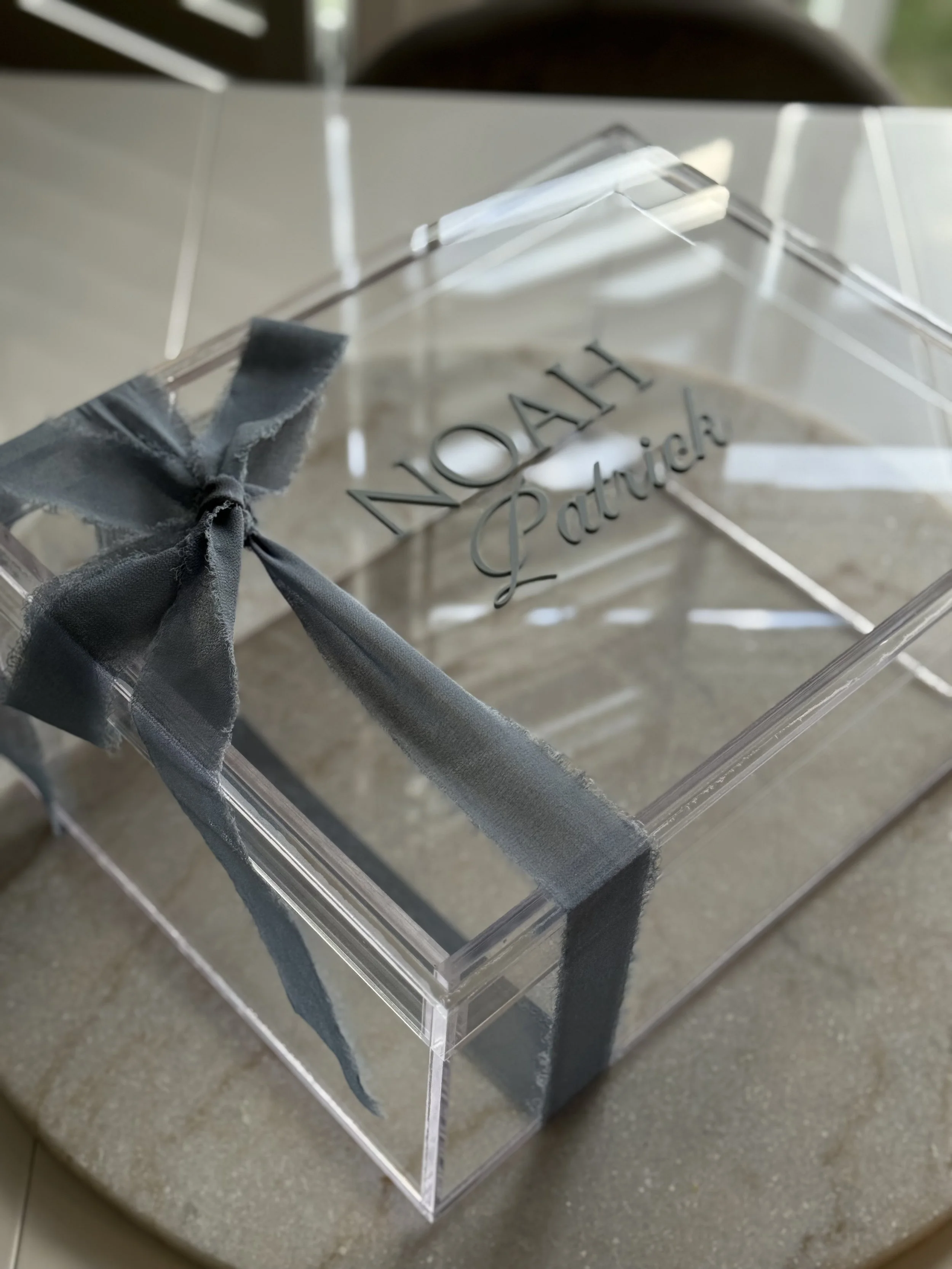A clear acrylic gift box with a silver ribbon bow on top, labeled with the names 'Hao' and 'Patricia' on the lid.