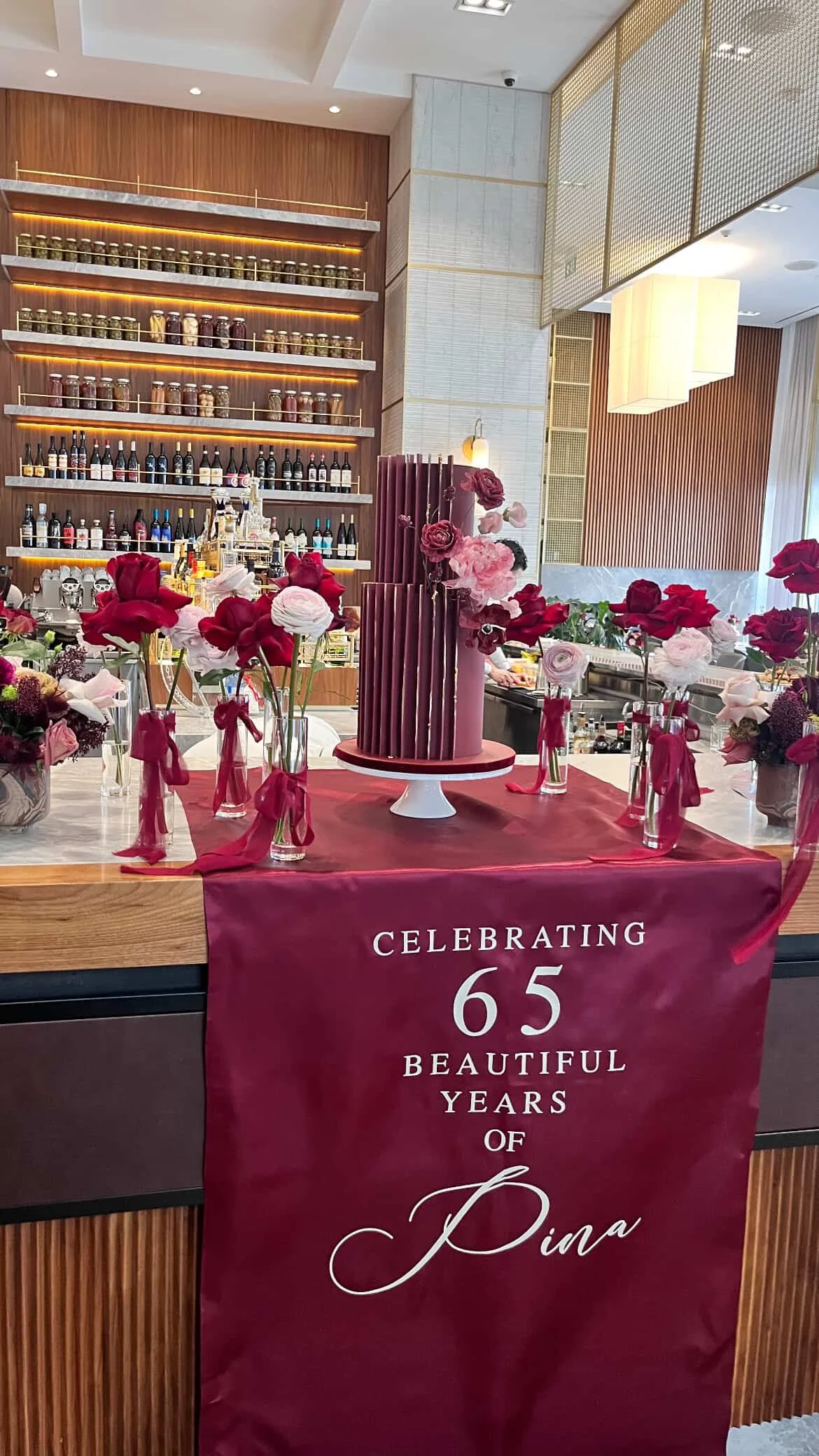 Decorated table with pink and red flowers celebrating Dina's 65th birthday, with a cake in the center and a bar in the background.