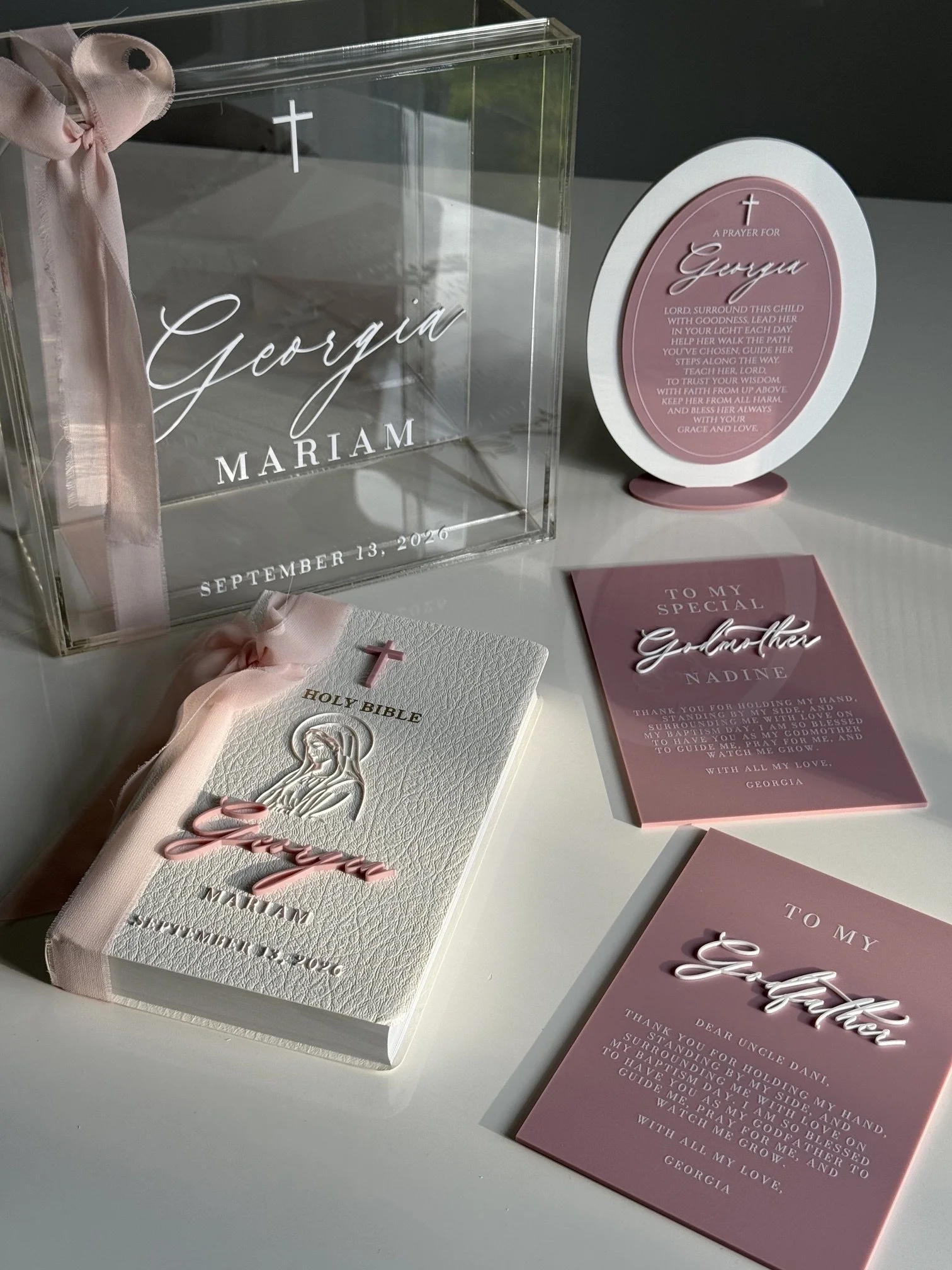 A gift box with a pink ribbon and cross, containing a white Orthodox Christian prayer book with pink accents, alongside pink prayer cards and a decorative sign expressing a prayer for a girl named Georgia.