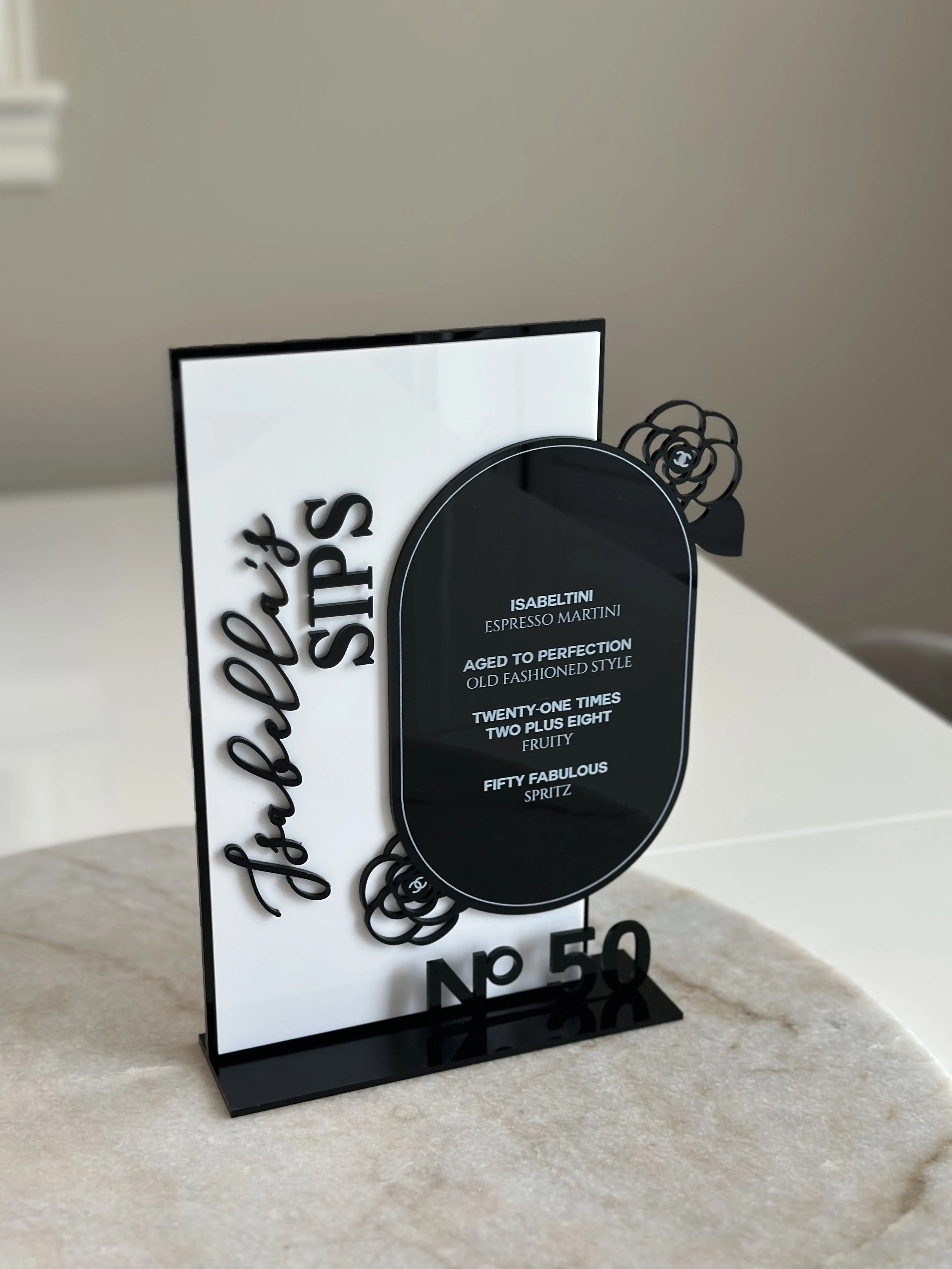 Decorative black and white sign with text and floral design elements, standing on a beige marble surface.