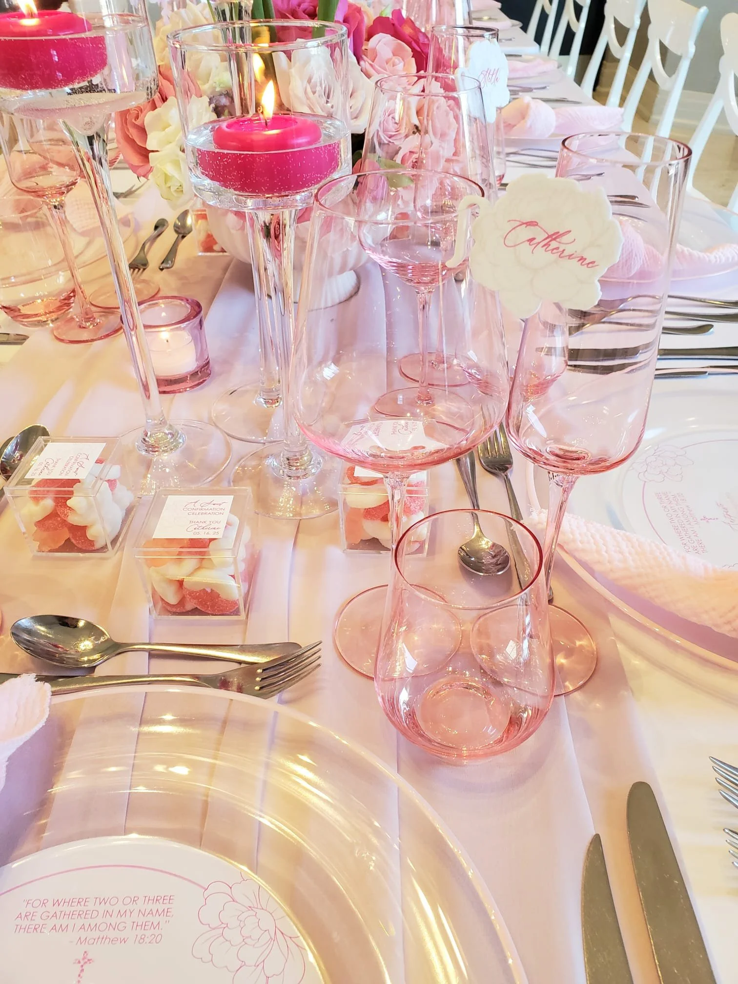 A pink-themed wedding or celebration table setup with a centerpiece, candles, and pink glassware, including wine glasses and a candle in a glass holder, with place cards, cutlery, and decorative elements.