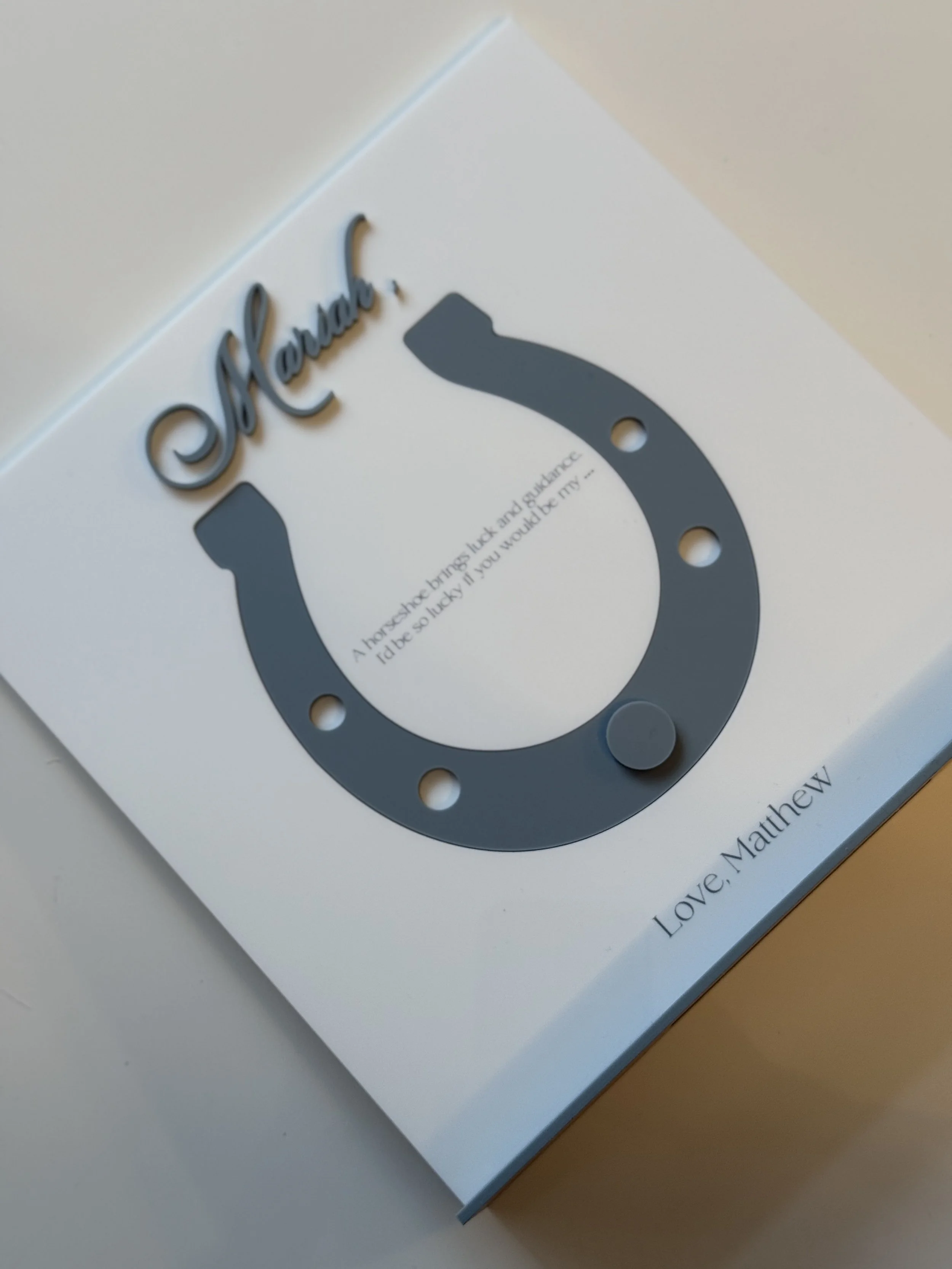 A white card with the word 'Mama' in cursive at the top, featuring a dark gray horseshoe-shaped decorative piece with a small round object attached, and a quote inside the horseshoe reading, 'A horseshoe brings luck and guidance. I'd be so lucky if y