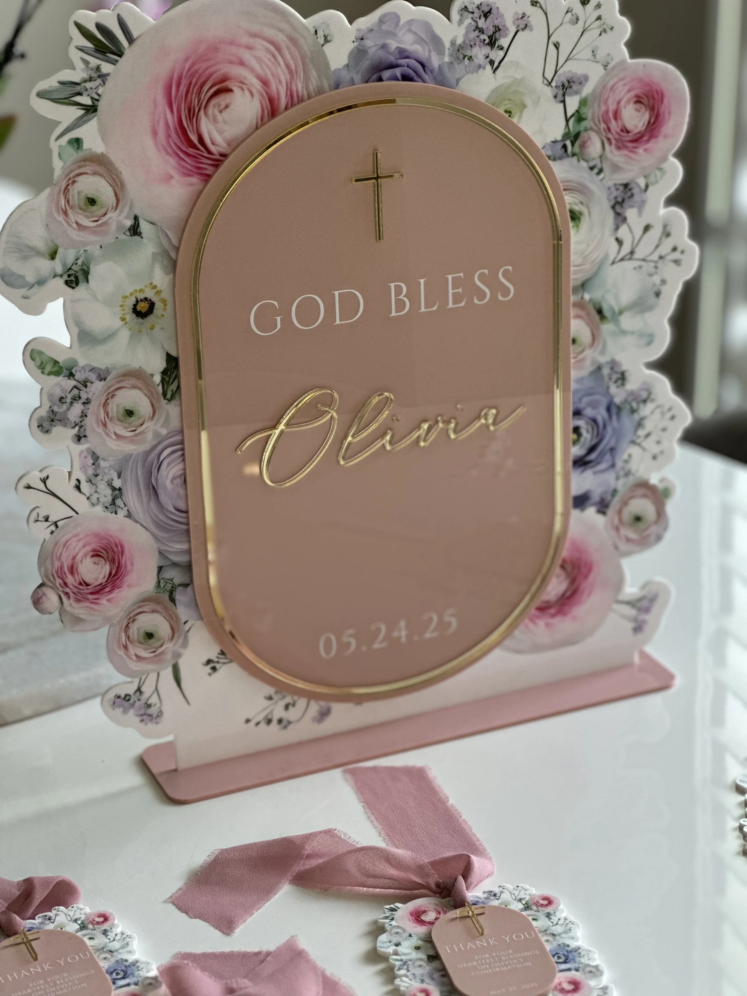 A pink floral decoration with a cross and the words "God Bless Olivia" along with a date, 05.24.25, on a pink oval plaque.