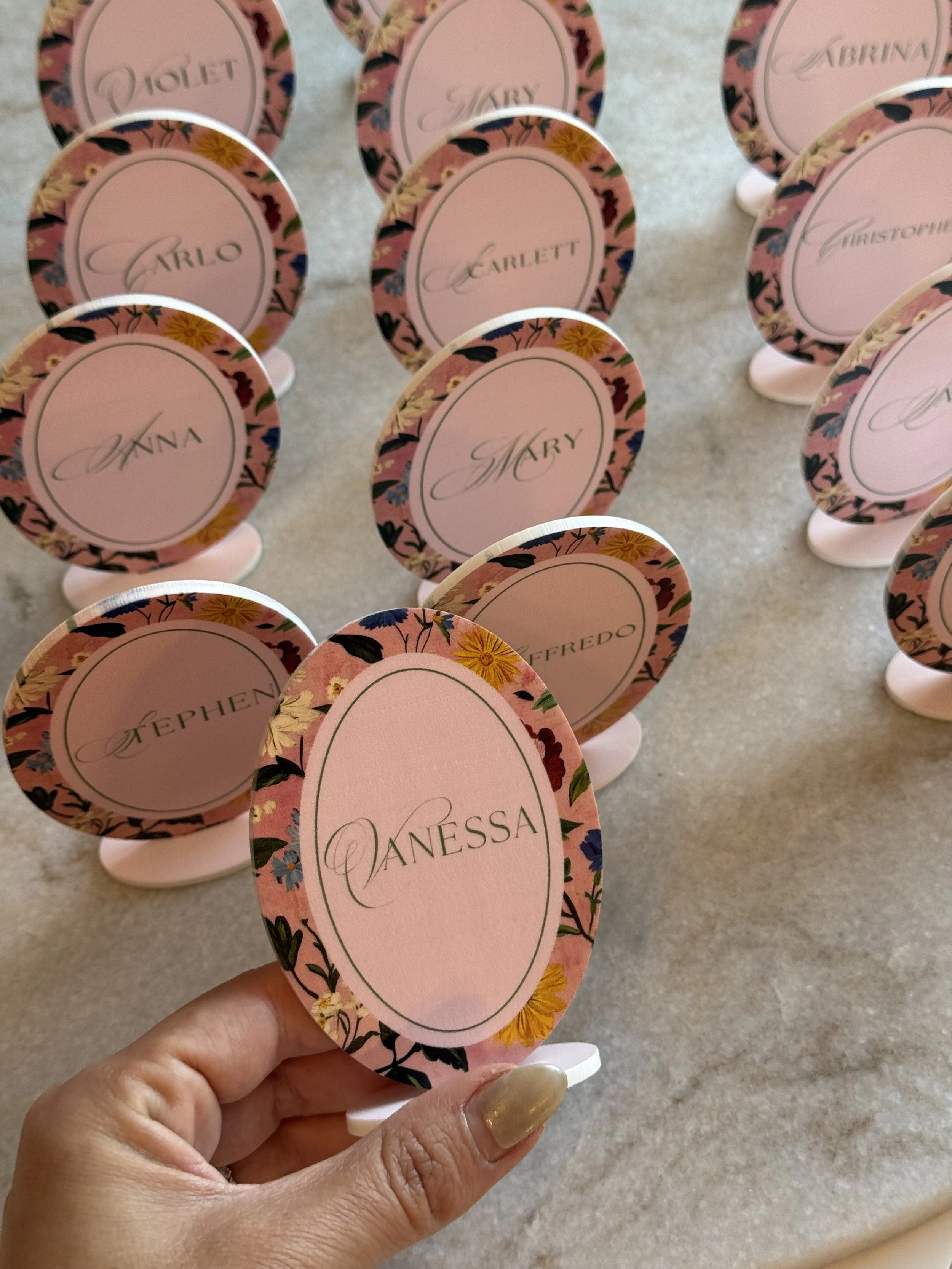 Decorative table markers with floral borders and the name 'Vanessa' written in elegant script, held by a hand with gold nail polish.