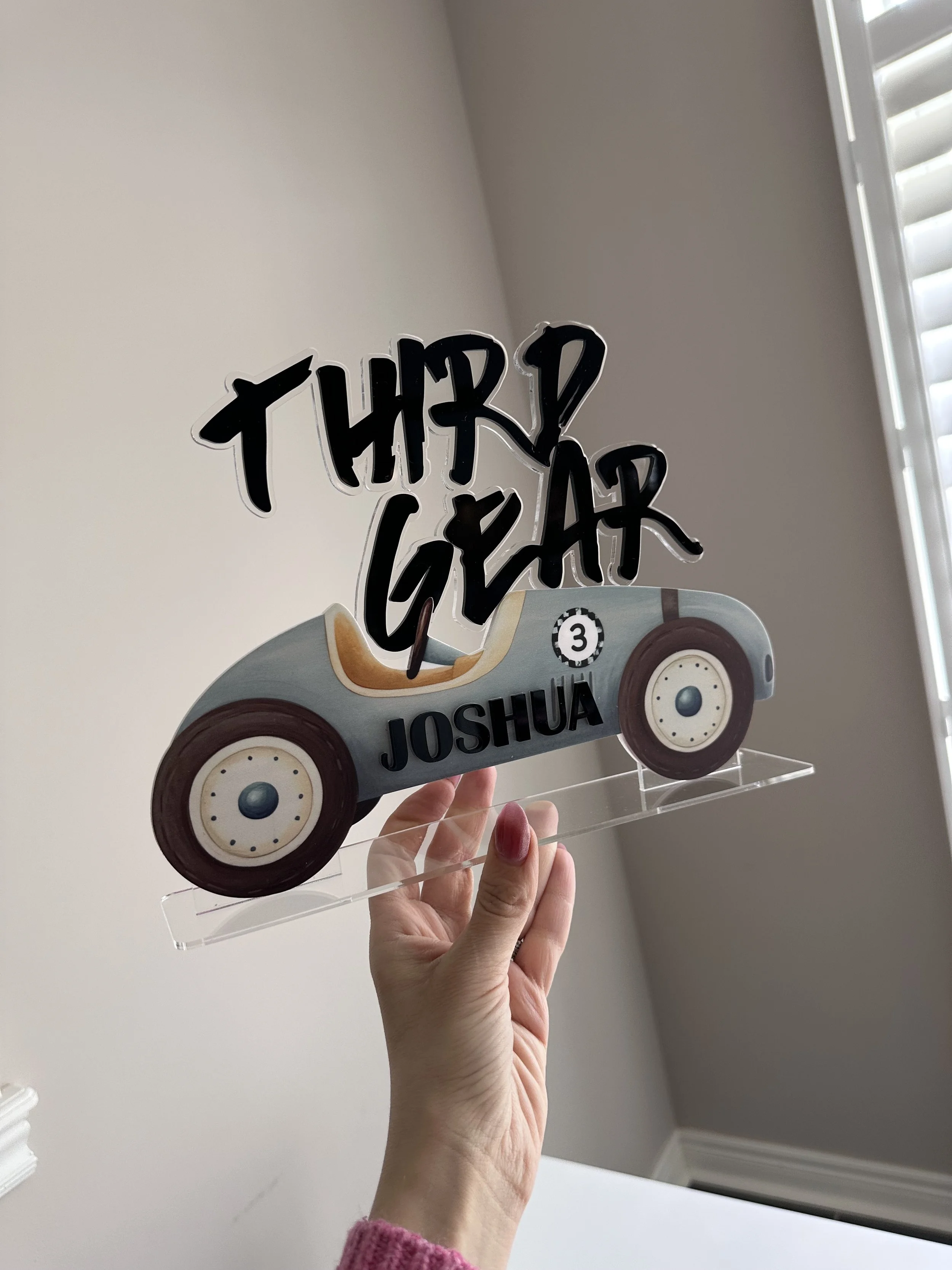 A decorative birthday sign with a race car design, inscribed with 'THUR! GEAR' on top, 'JOSHUA' at the bottom, and a small number 3 in a circle.