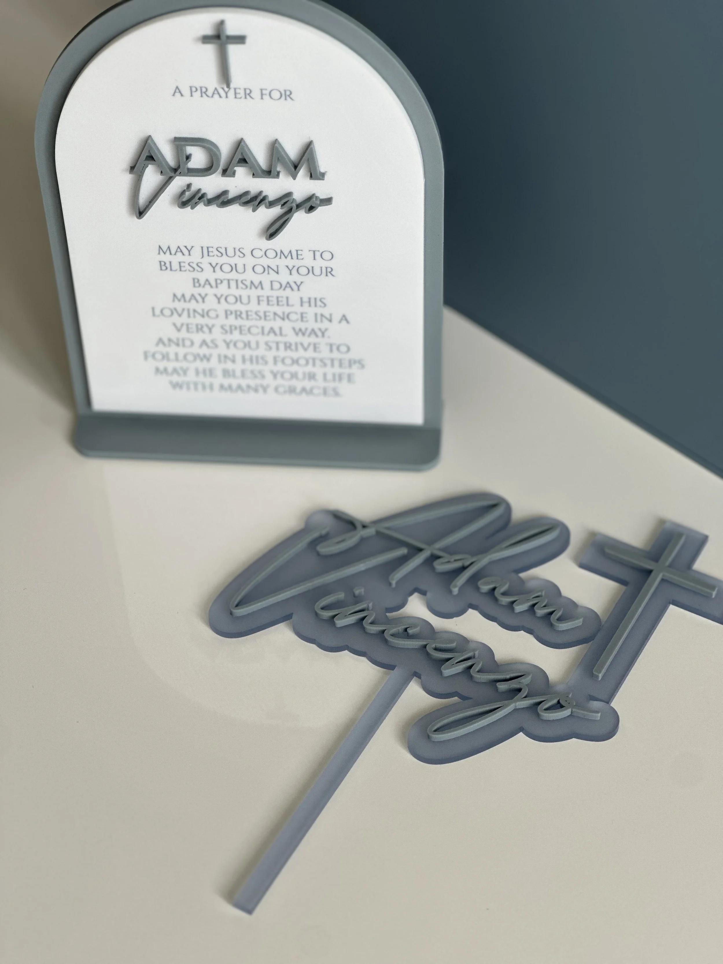 Decorative prayer sign with a cross and text for Adam Vincenzo's baptism, and a cake topper with the word "Baptism" and a cross.