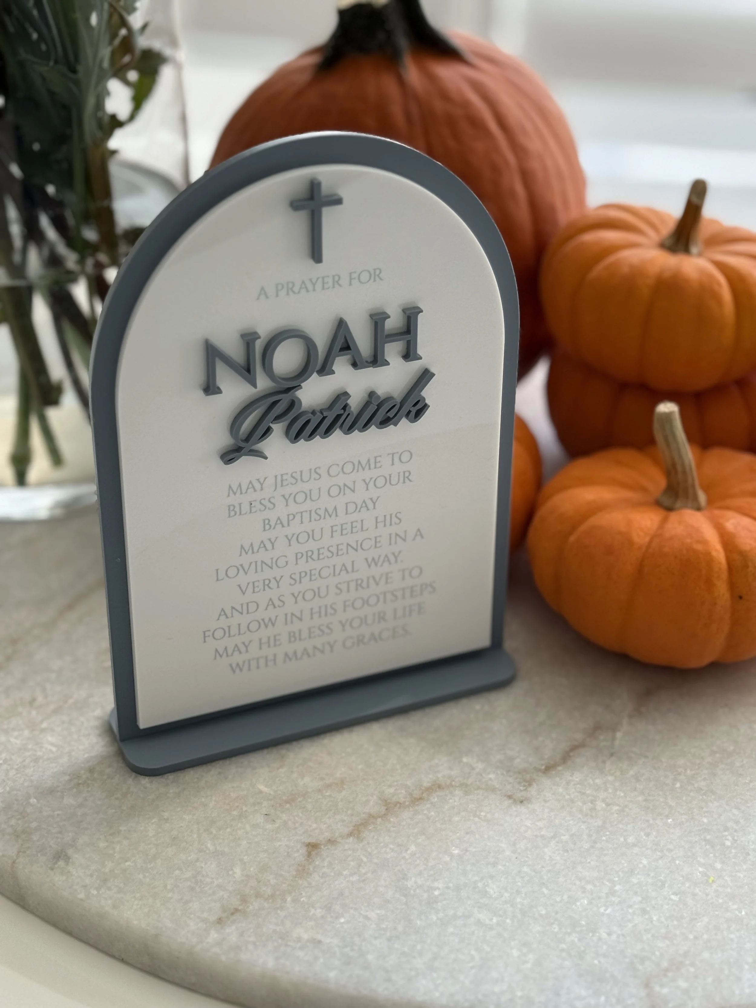A decorative plaque with a prayer for Noah Patrick, feature a cross at the top, placed on a white marble surface, with small orange pumpkins around it.