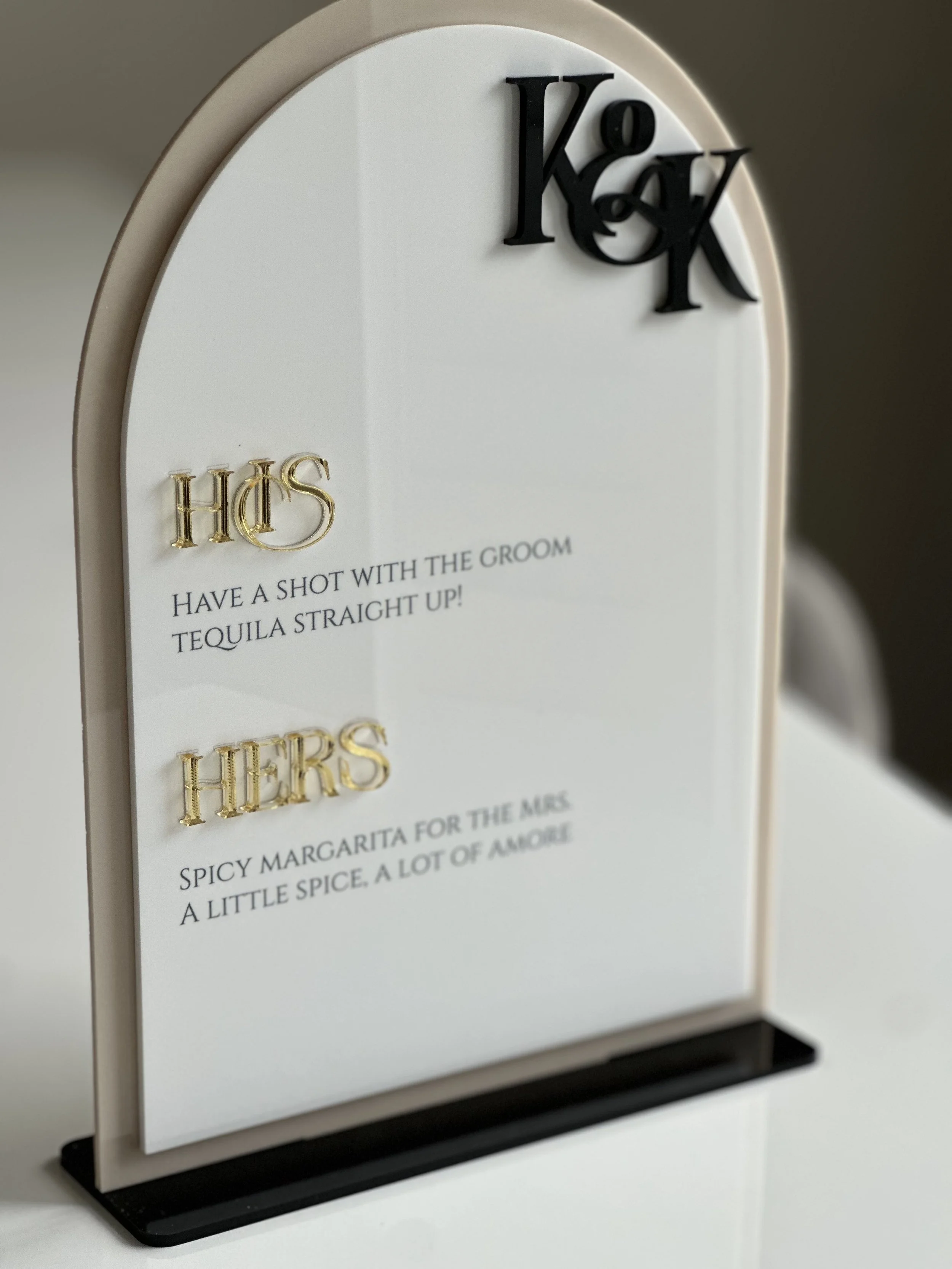 Wedding table sign with black and gold lettering, displaying the words 'K & K' at the top, and two humorous messages about wedding toasts and margaritas.