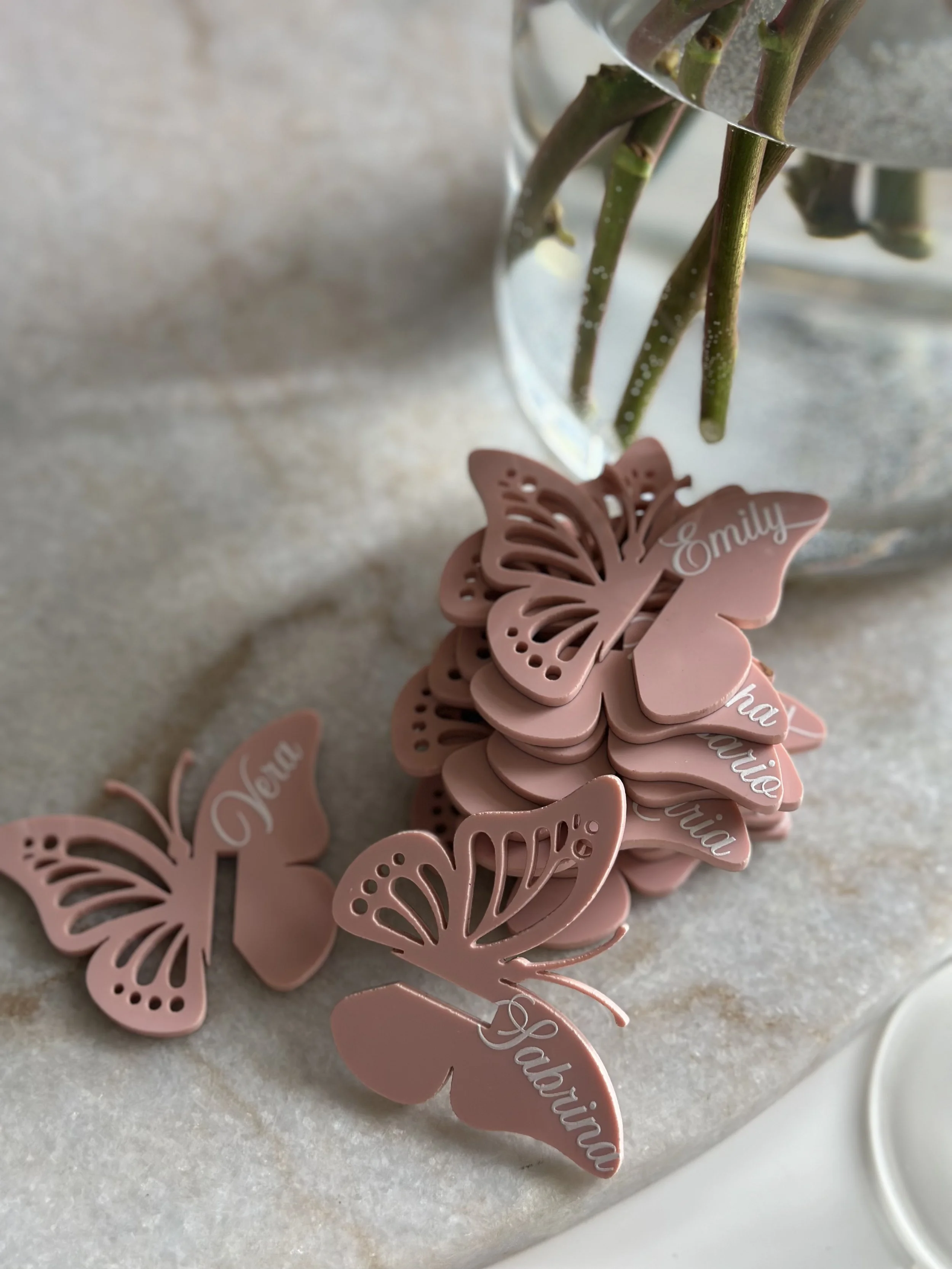 Pink butterfly-shaped tags with white cursive writing of names, arranged on a light-colored surface near a glass vase with stems of greenery.